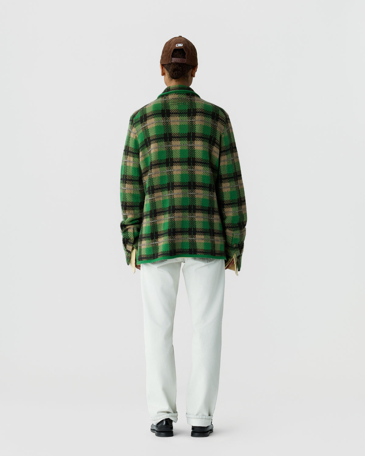 Person wearing a green and brown plaid jacket, white pants, black shoes, and a brown cap, standing facing away against a plain white background.