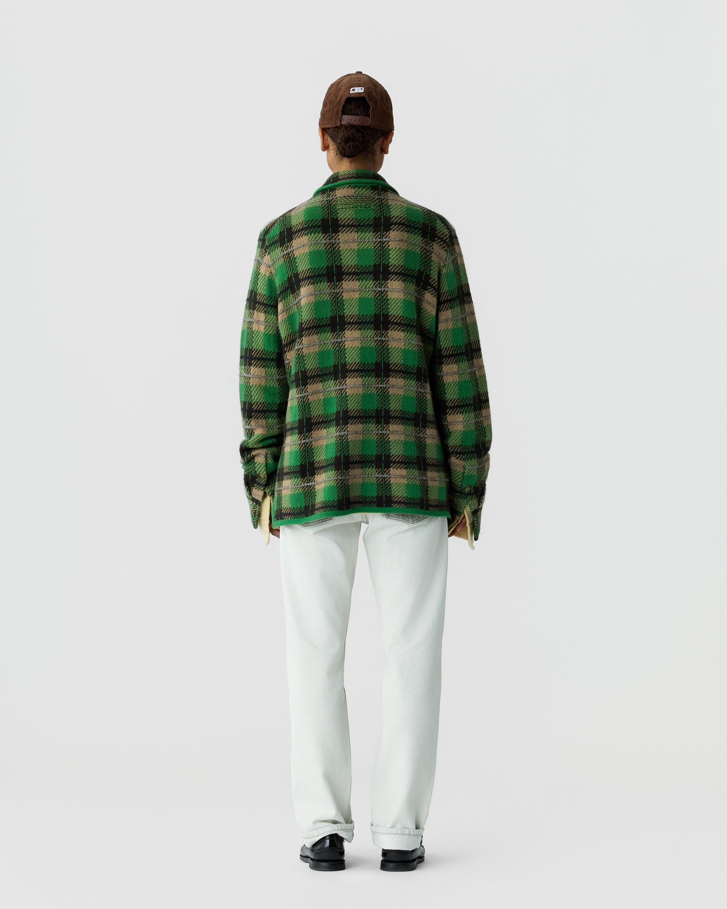 Person wearing a green and brown plaid jacket, white pants, black shoes, and a brown cap, standing facing away against a plain white background.