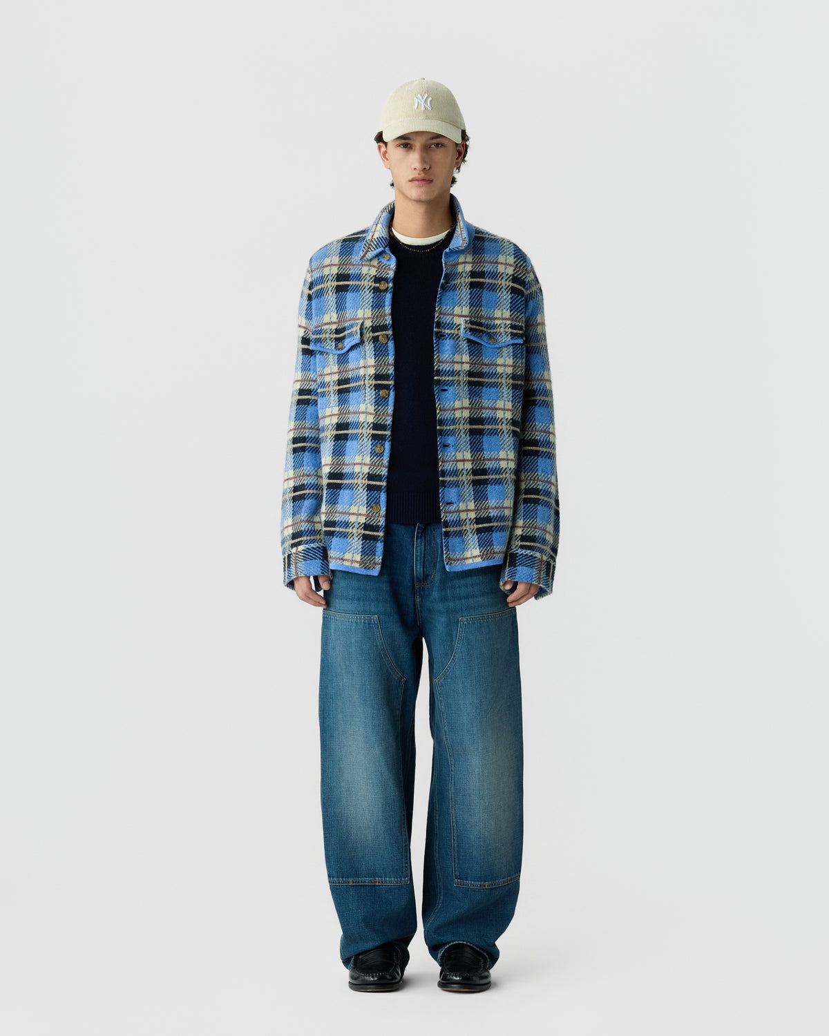 A man wearing a beige baseball cap, a blue and yellow plaid button-up shirt over a dark sweater, loose-fitting blue jeans, and black shoes standing against a plain light background.