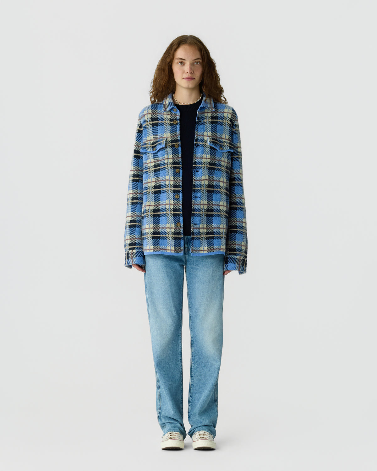 Young woman with curly brown hair wearing a blue and beige plaid button-up shirt over a black top, light blue jeans, and white sneakers, standing against a plain white background.