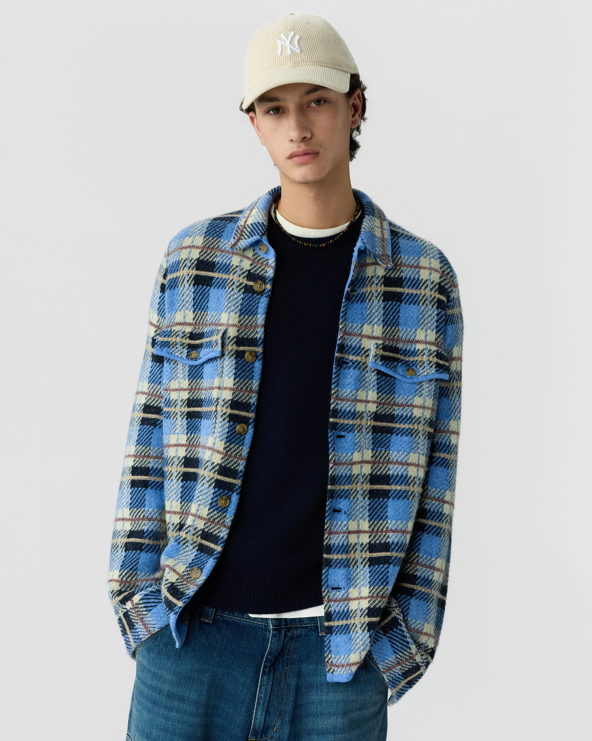 Young man wearing a beige baseball cap, navy sweater, and a blue and beige plaid button-up shirt over blue jeans against a plain light gray background.