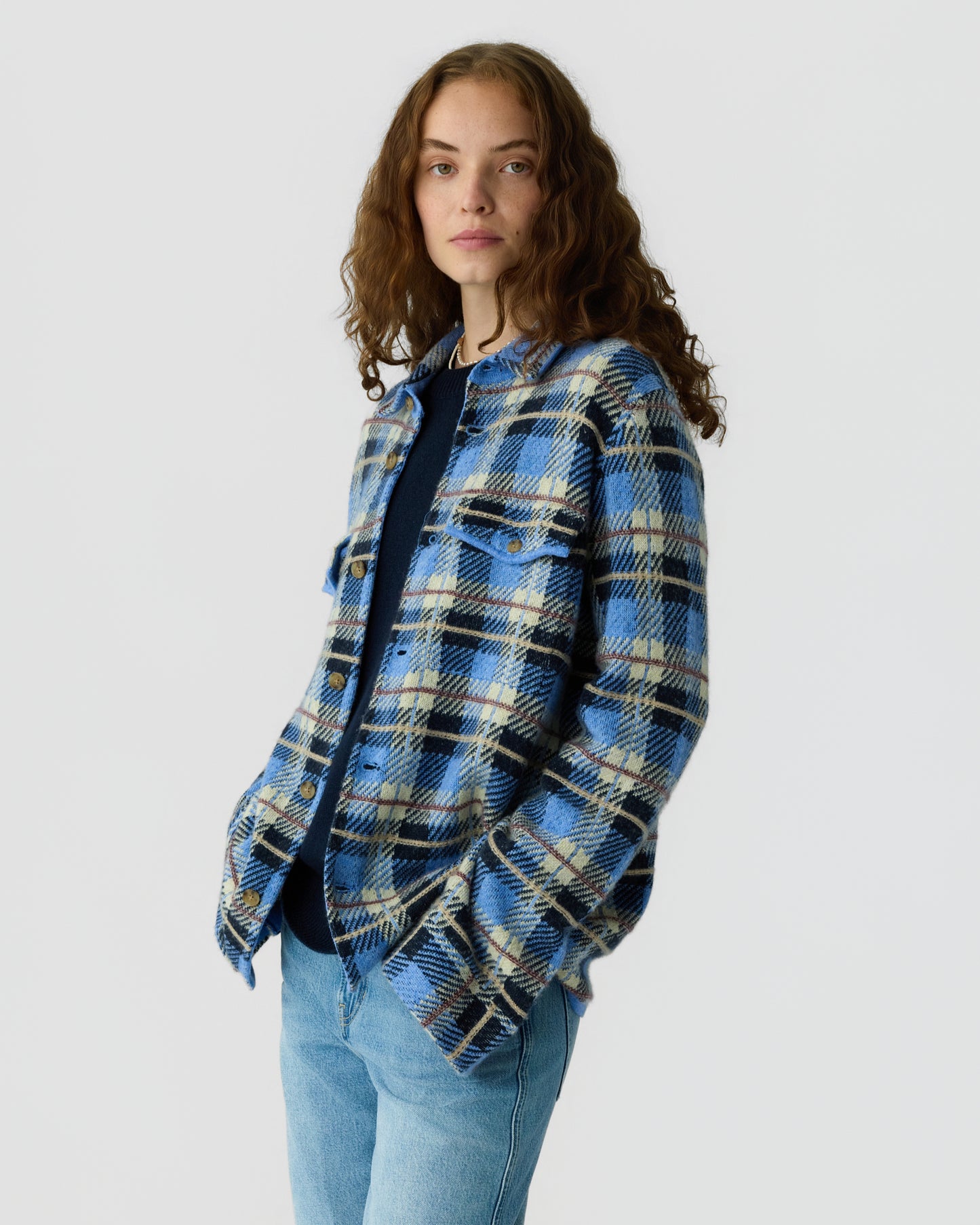 Young woman with curly brown hair wearing a blue, cream, and black plaid jacket over a dark top and light blue jeans, standing against a plain white background.