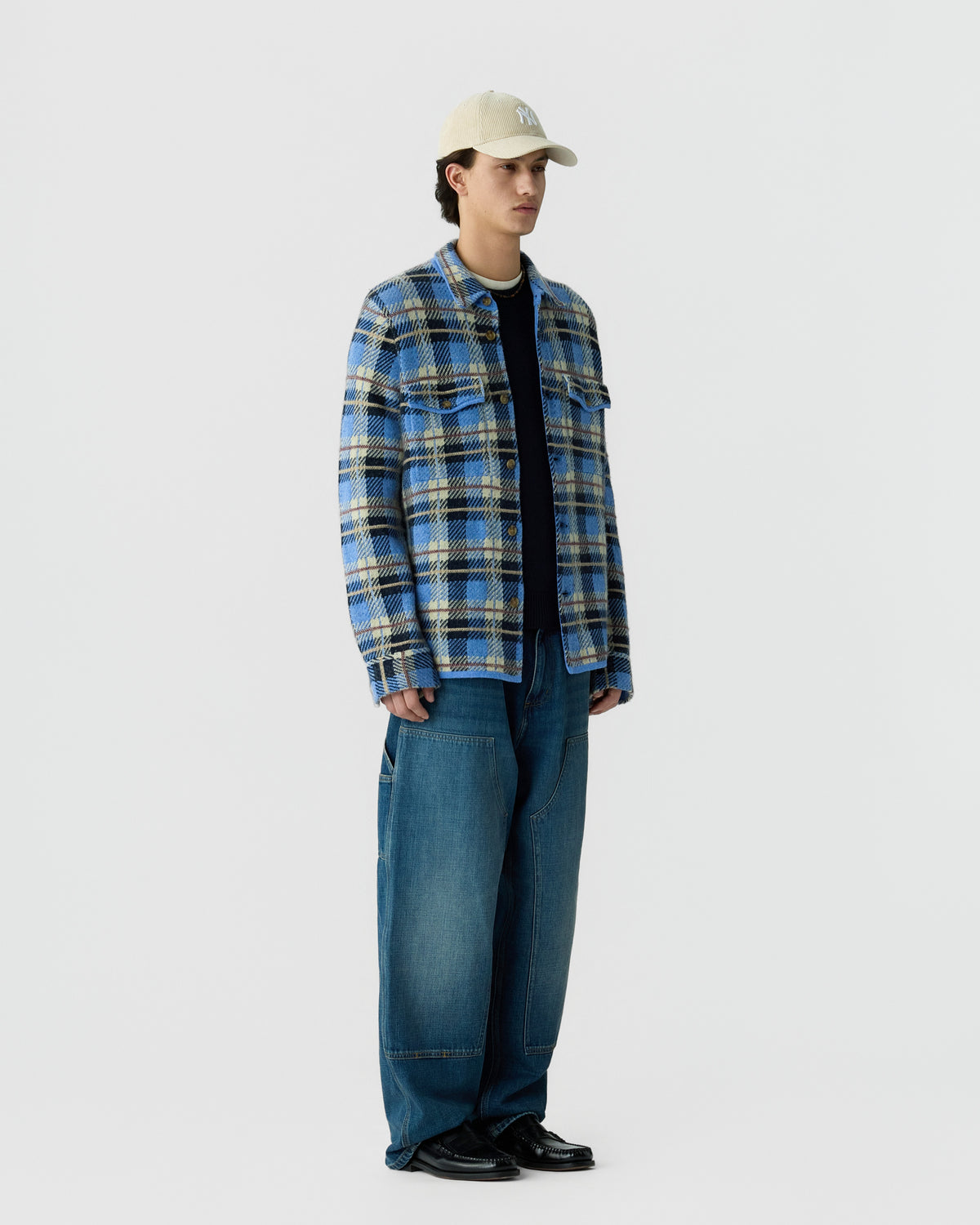 A young man stands against a plain light background wearing a blue and beige plaid jacket over a black shirt, loose blue jeans, black shoes, and a beige baseball cap.