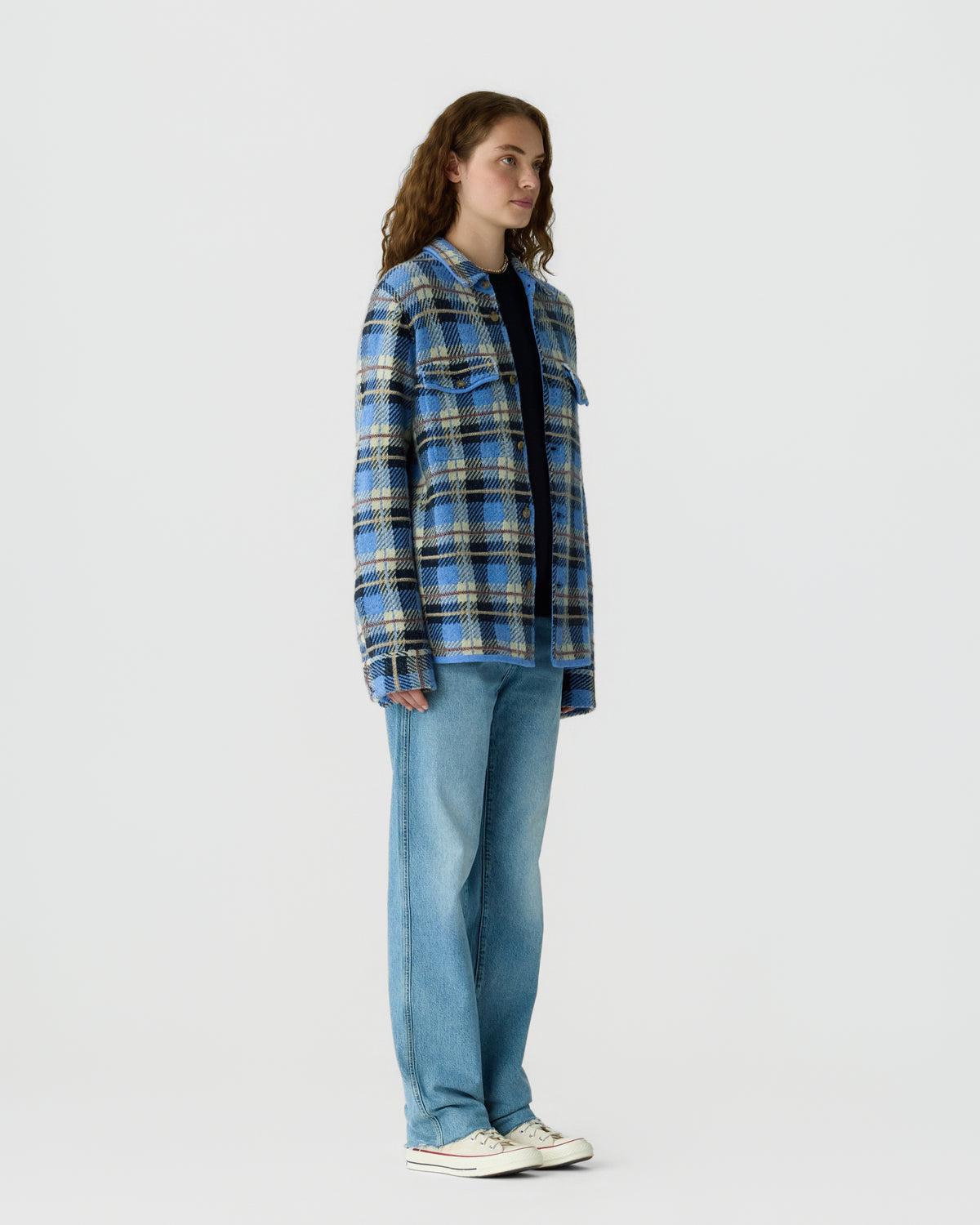 A woman stands facing sideways wearing a loose blue, black, and white plaid jacket over a dark shirt, paired with light blue wide-leg jeans and white sneakers against a plain background.