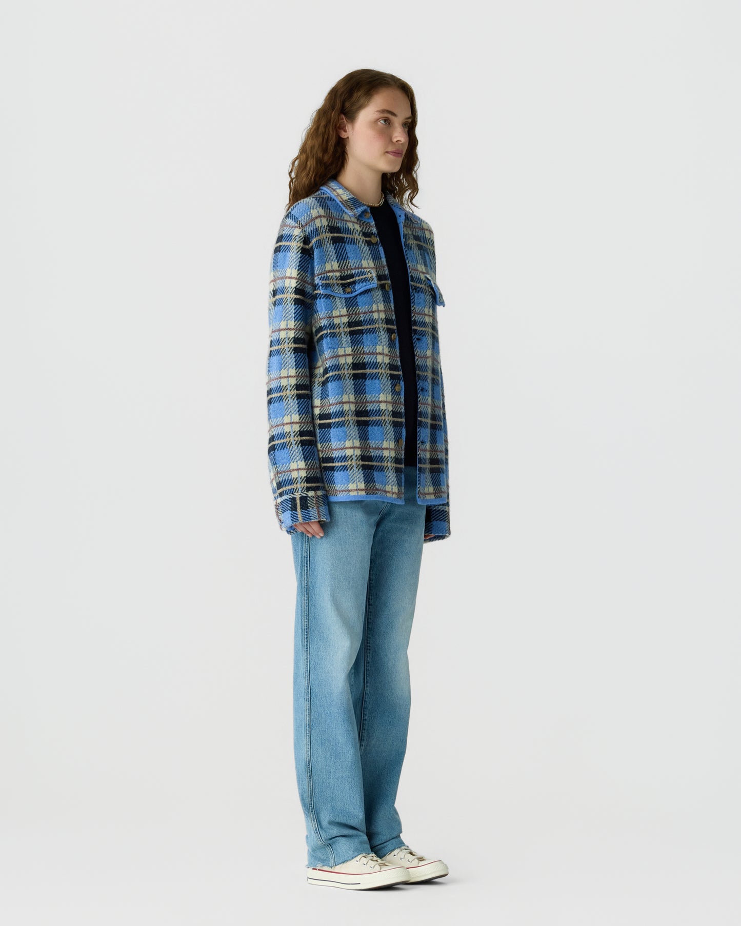 A woman stands facing sideways wearing a loose blue, black, and white plaid jacket over a dark shirt, paired with light blue wide-leg jeans and white sneakers against a plain background.