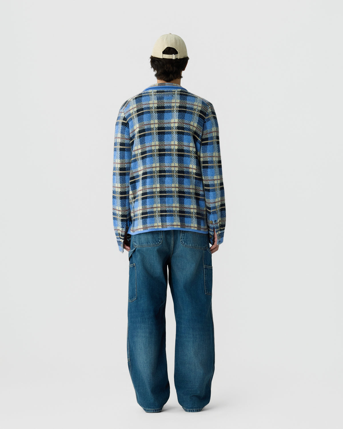 Person wearing a blue and beige plaid long-sleeve shirt, baggy blue jeans, and a light beige cap, standing facing away against a plain white background.