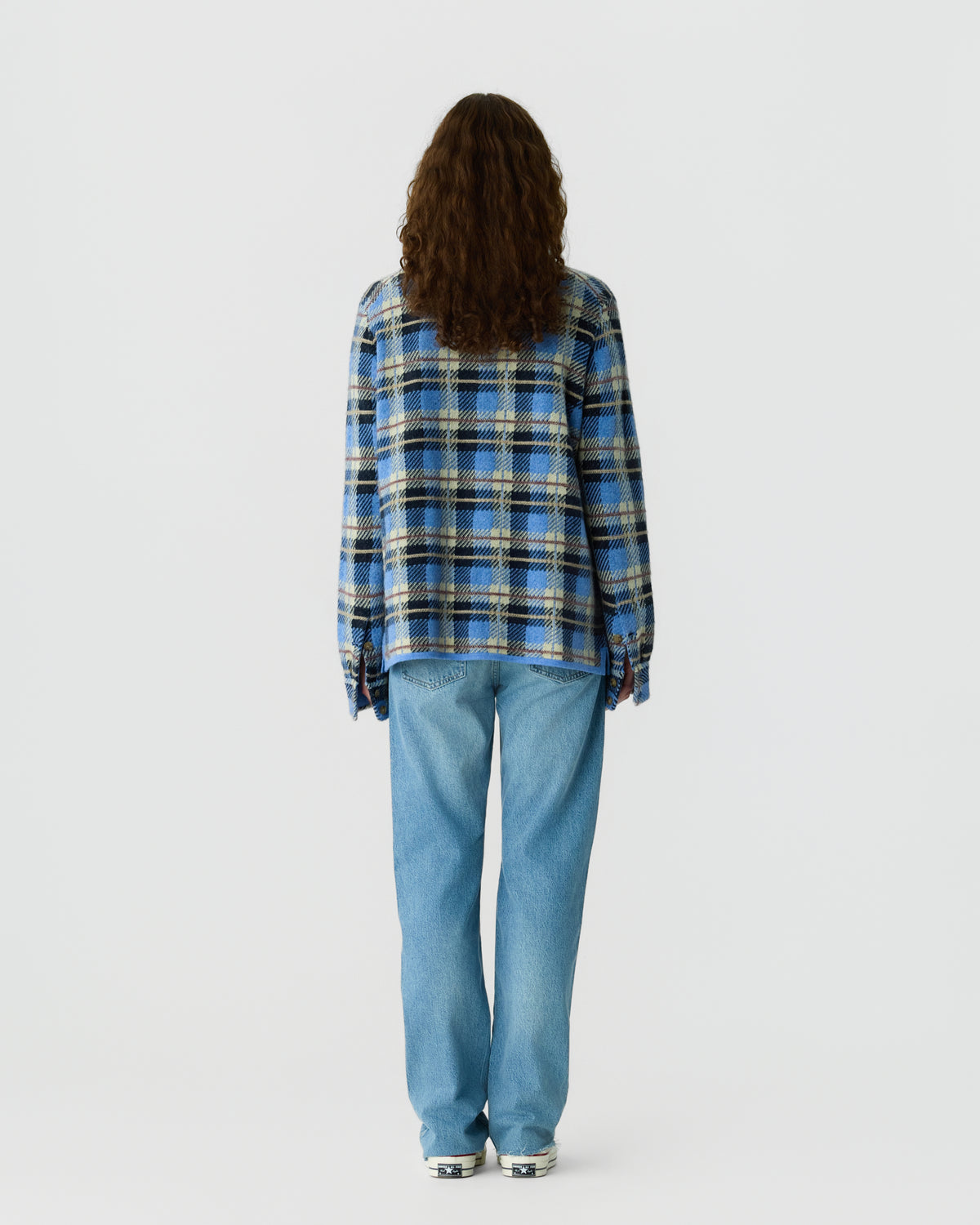 Person with long curly hair seen from behind wearing a blue, black, and tan plaid shirt paired with light blue jeans and white sneakers against a plain background.