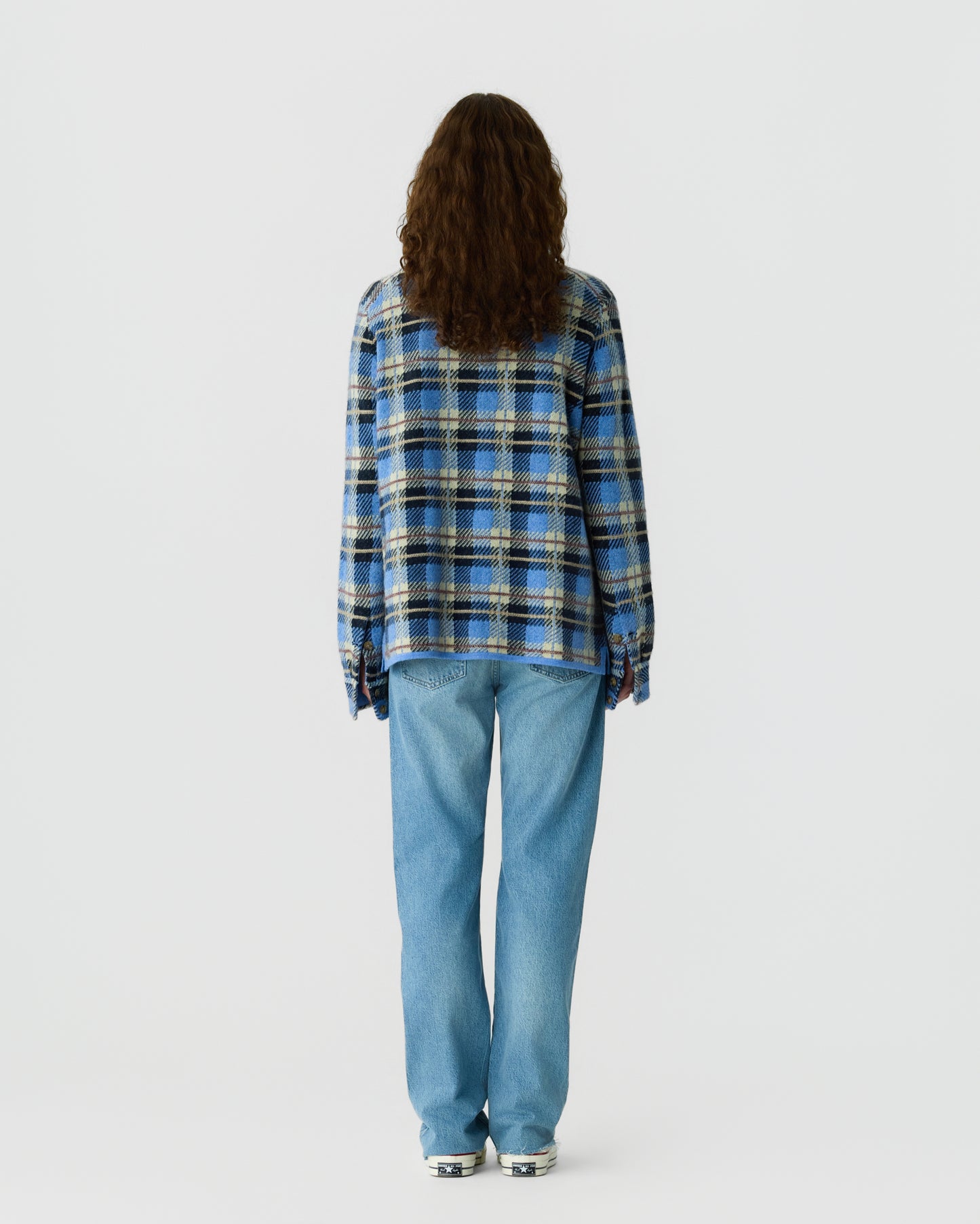 Person with long curly hair seen from behind wearing a blue, black, and tan plaid shirt paired with light blue jeans and white sneakers against a plain background.