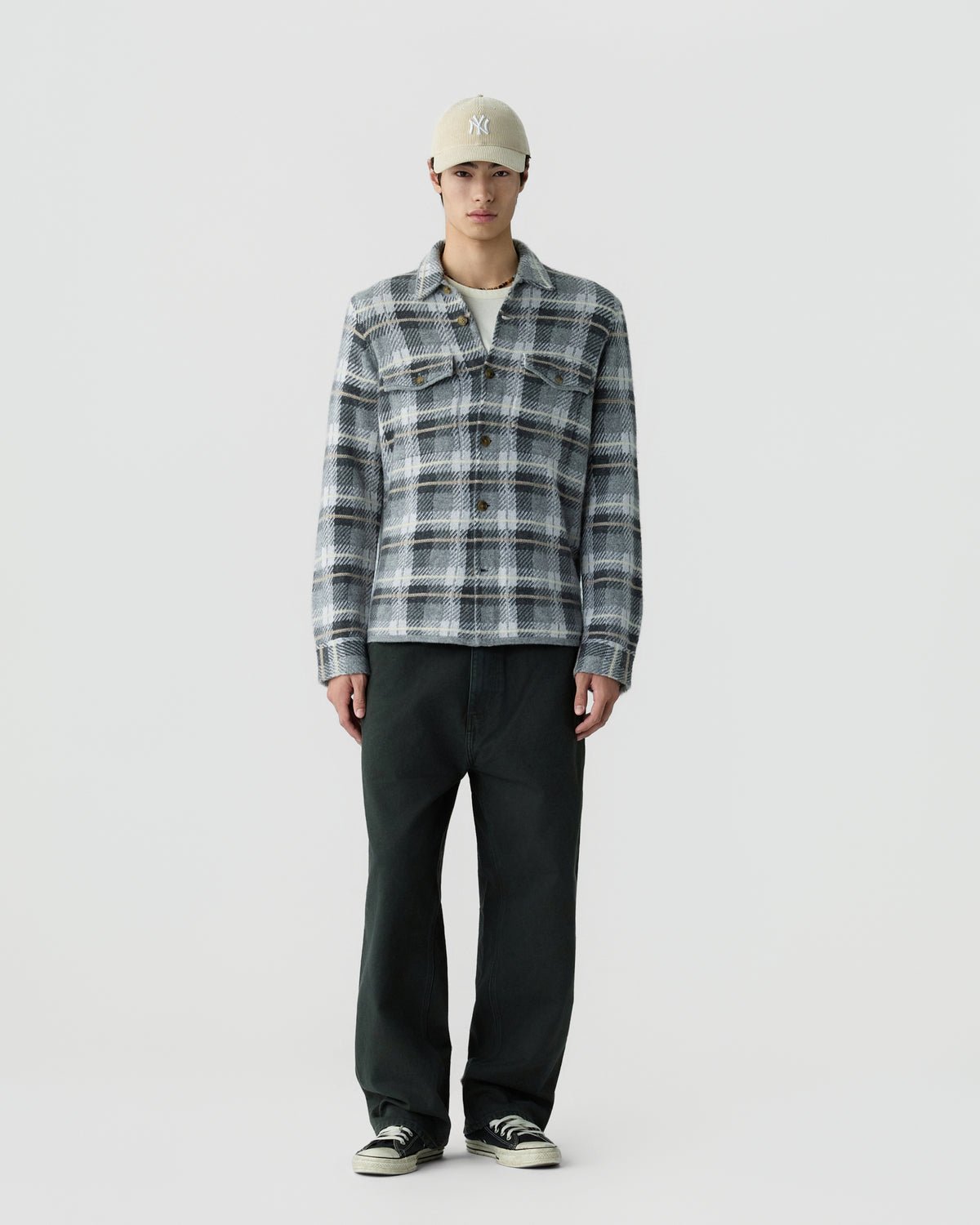 A man stands against a plain background wearing a beige baseball cap, a gray and white plaid long-sleeve shirt jacket with buttons, loose black pants, and black and white sneakers.