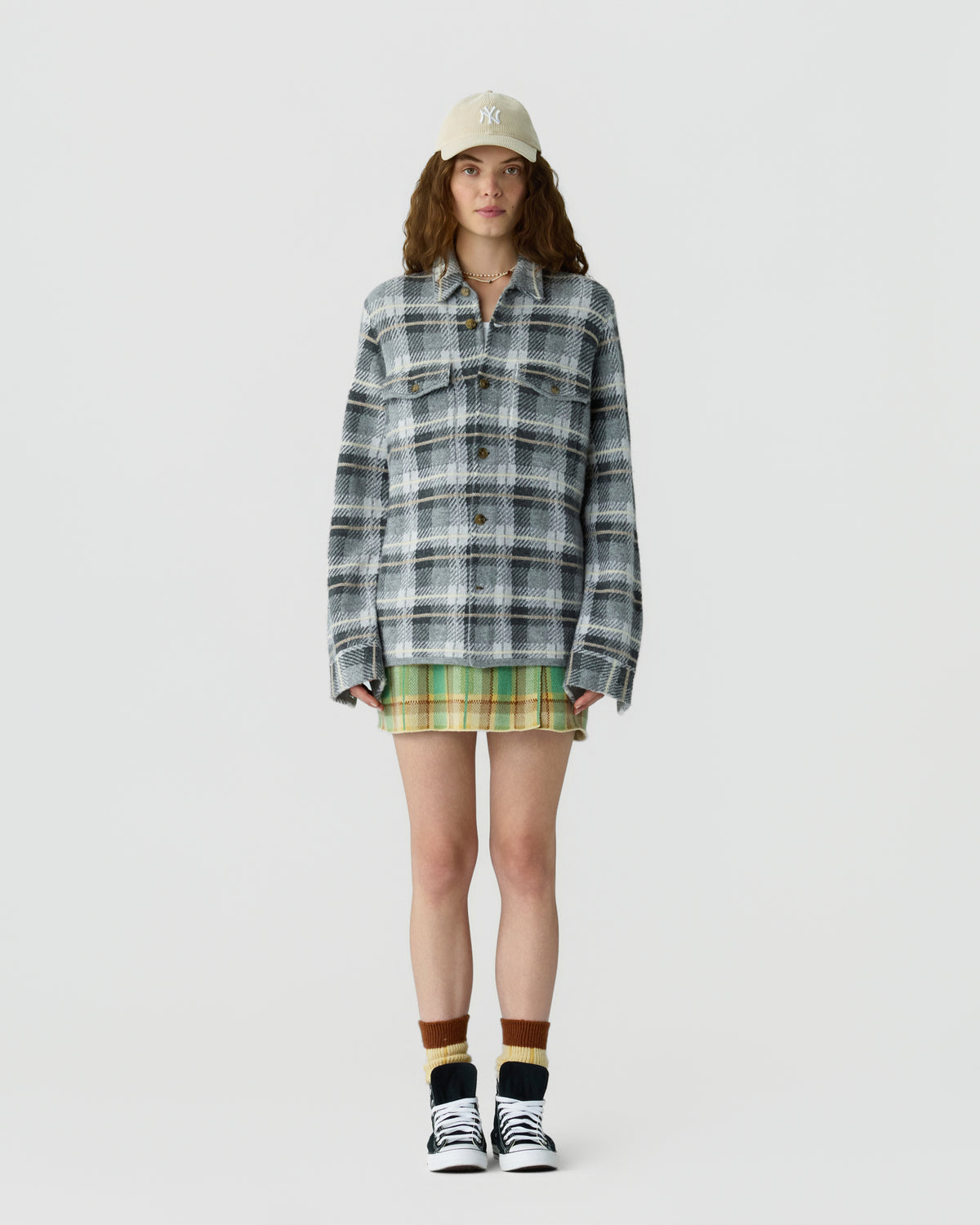 Young woman wearing a beige baseball cap, oversized gray and white plaid jacket, green plaid skirt, brown and beige socks, and black and white sneakers standing against a plain white background.
