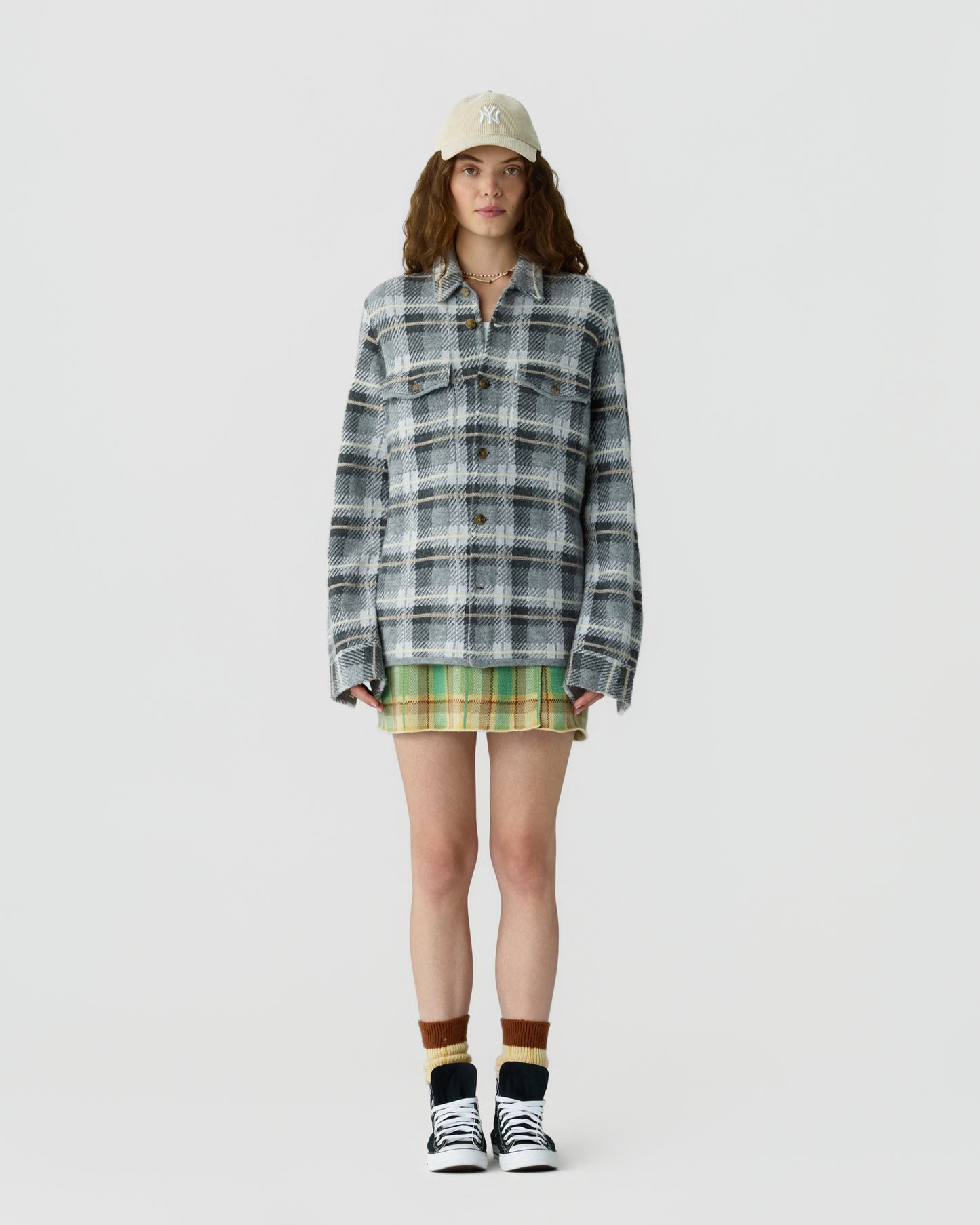 Young woman wearing a beige baseball cap, oversized gray and white plaid jacket, green plaid skirt, brown and beige socks, and black and white sneakers standing against a plain white background.