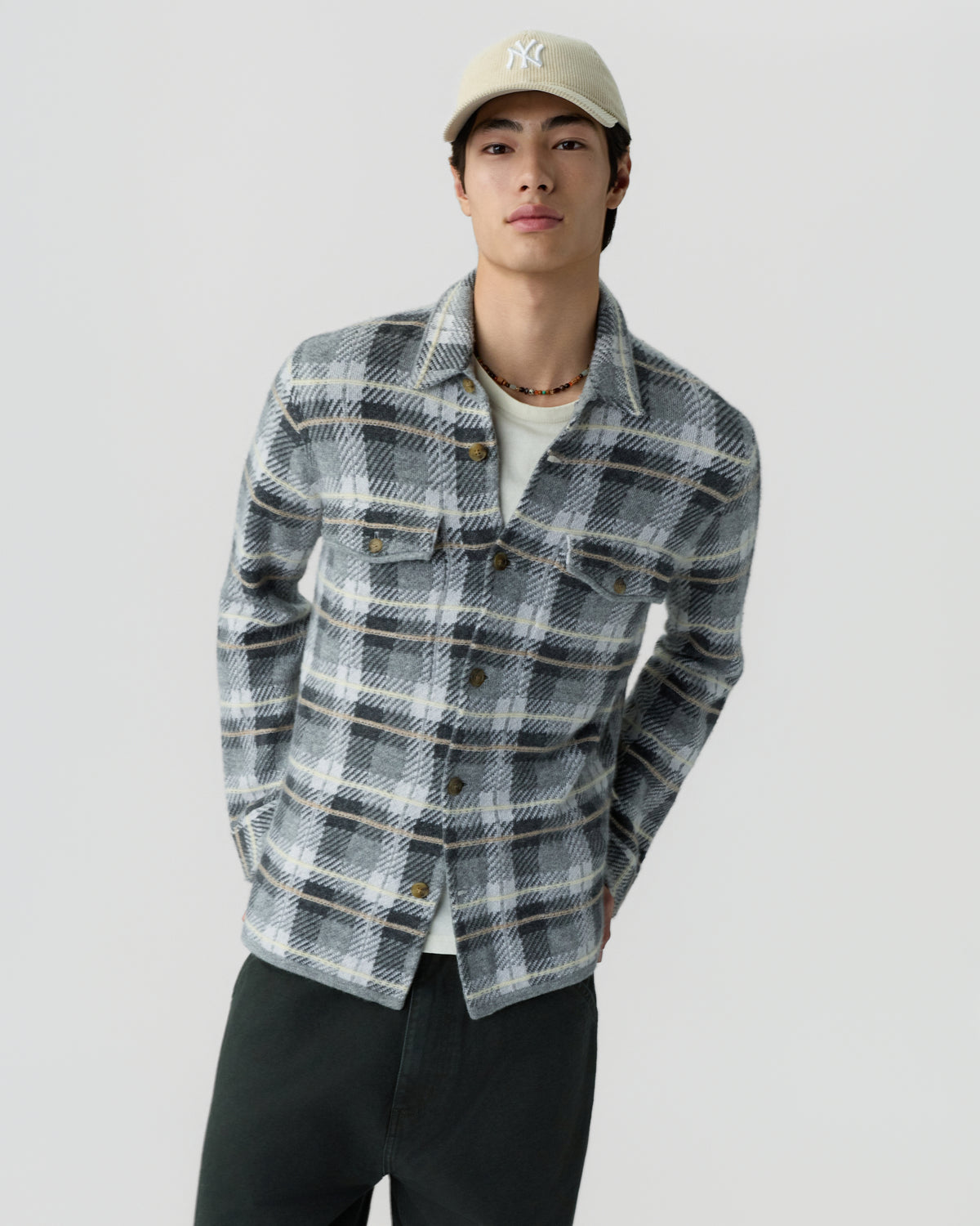 Young man wearing a gray and white plaid button-up shirt over a white t-shirt, black pants, and a beige baseball cap against a plain light background.