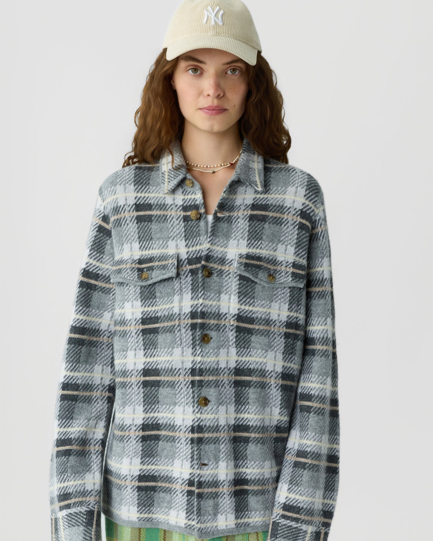 Young woman wearing a beige baseball cap and a gray and black plaid button-up shirt with chest pockets against a plain white background.