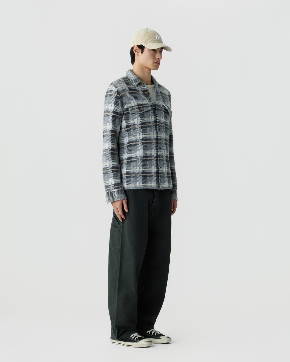 Young man wearing a gray and white plaid jacket, black wide-leg pants, black and white sneakers, and a beige baseball cap standing against a plain white background.