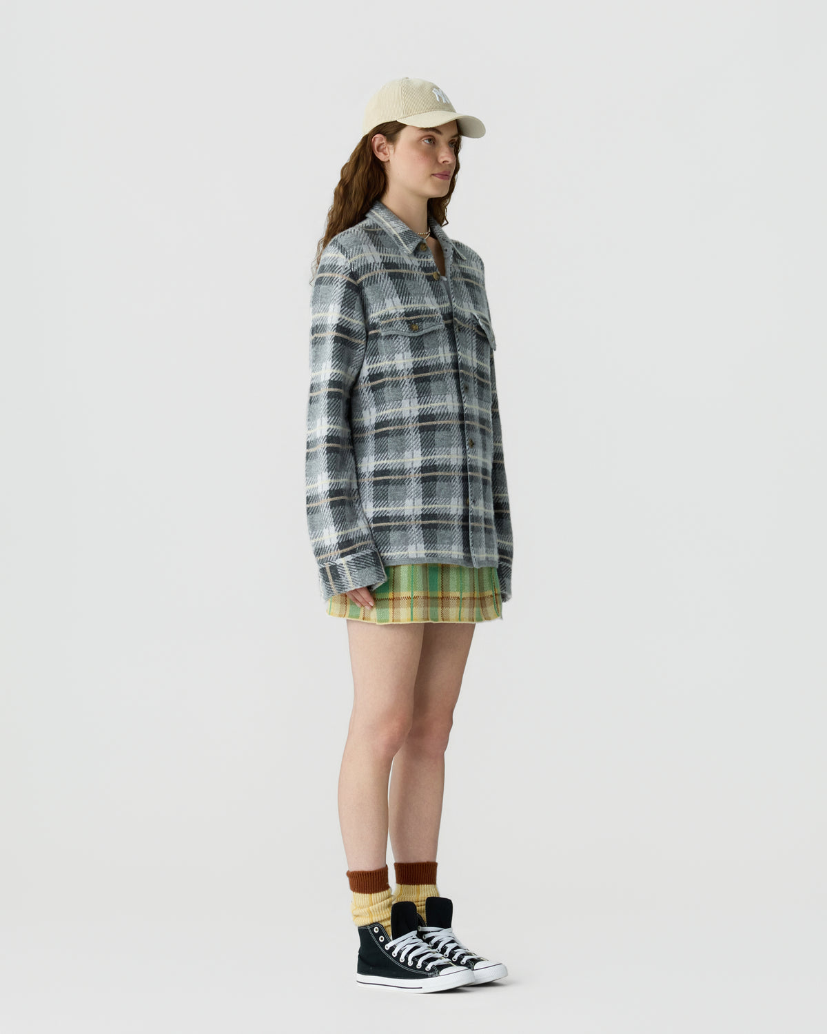 Woman wearing a beige cap, oversized blue and gray plaid jacket, green and yellow plaid skirt, striped socks, and black high-top sneakers standing against a plain white background.
