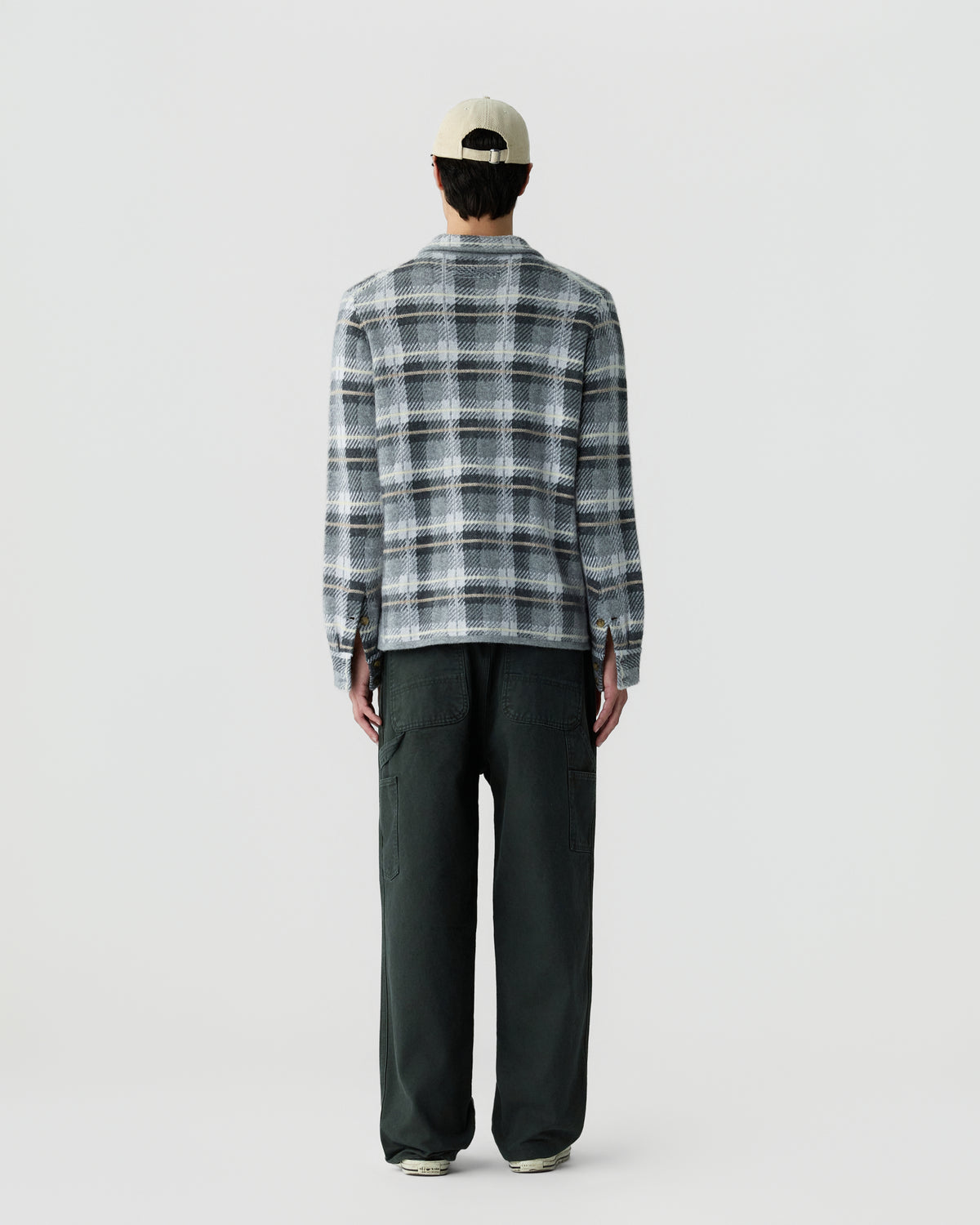Person wearing a beige cap, gray and white plaid long-sleeve shirt, and loose dark green pants, standing facing away against a plain white background.