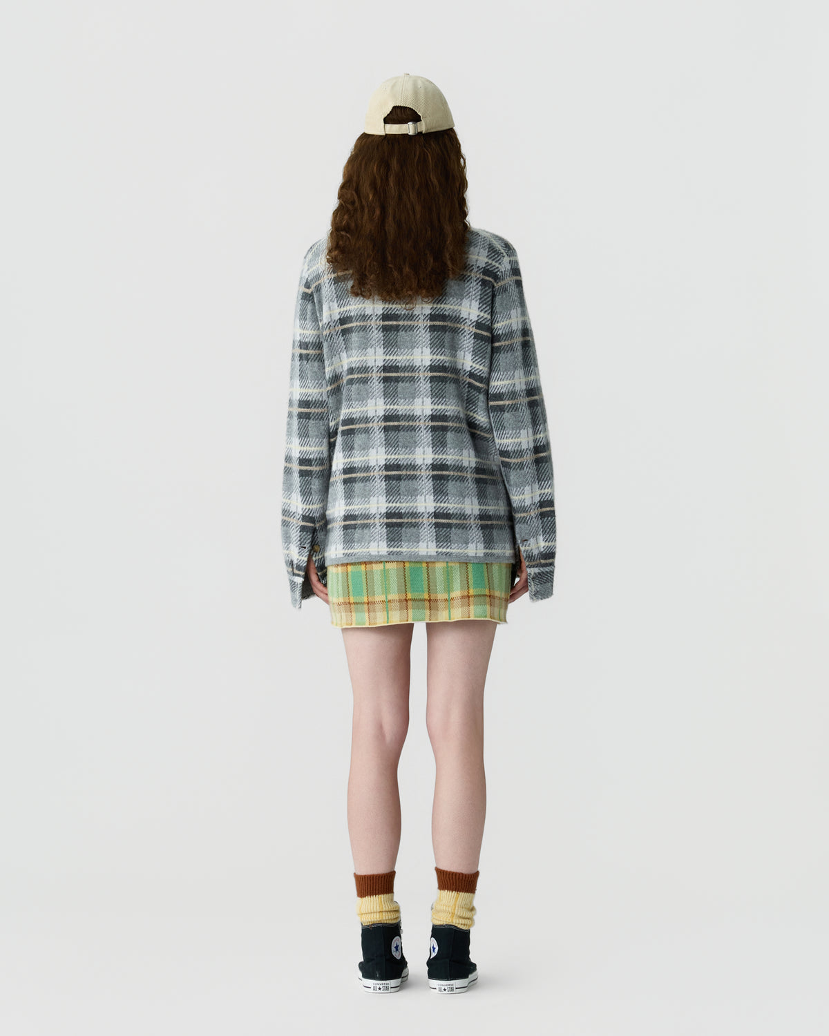 Person wearing a beige cap, long-sleeve gray and white plaid top, green and yellow plaid skirt, paired with yellow and brown socks and black sneakers, standing facing away against a plain background.