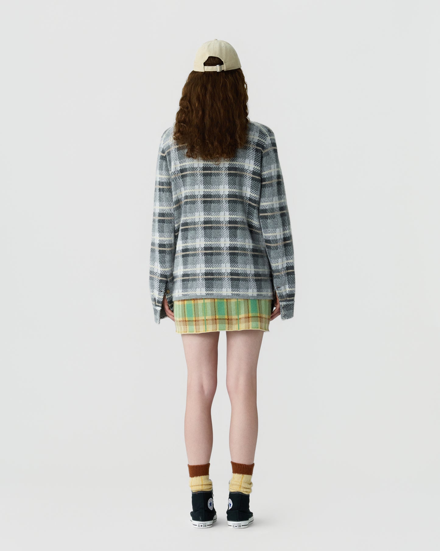 Person wearing a beige cap, long-sleeve gray and white plaid top, green and yellow plaid skirt, paired with yellow and brown socks and black sneakers, standing facing away against a plain background.