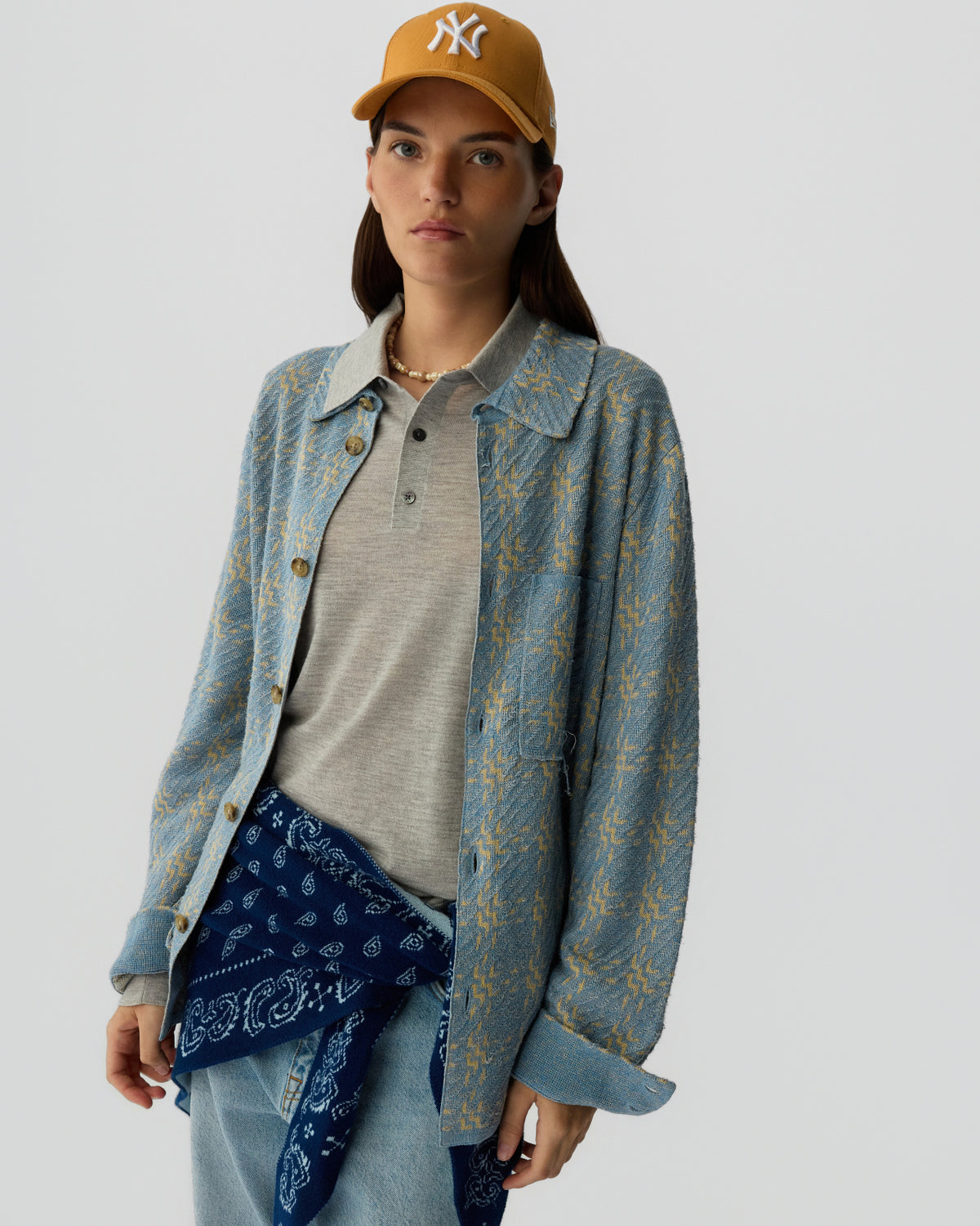 Woman wearing a mustard baseball cap with a light gray polo shirt under a blue patterned button-up shirt, light wash jeans, and a navy bandana tied around her waist against a plain gray background.