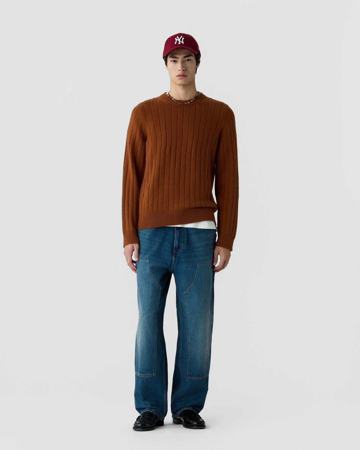 A young man wearing a brown ribbed sweater, blue relaxed-fit jeans, a white undershirt, black shoes, and a red baseball cap standing against a plain light gray background.