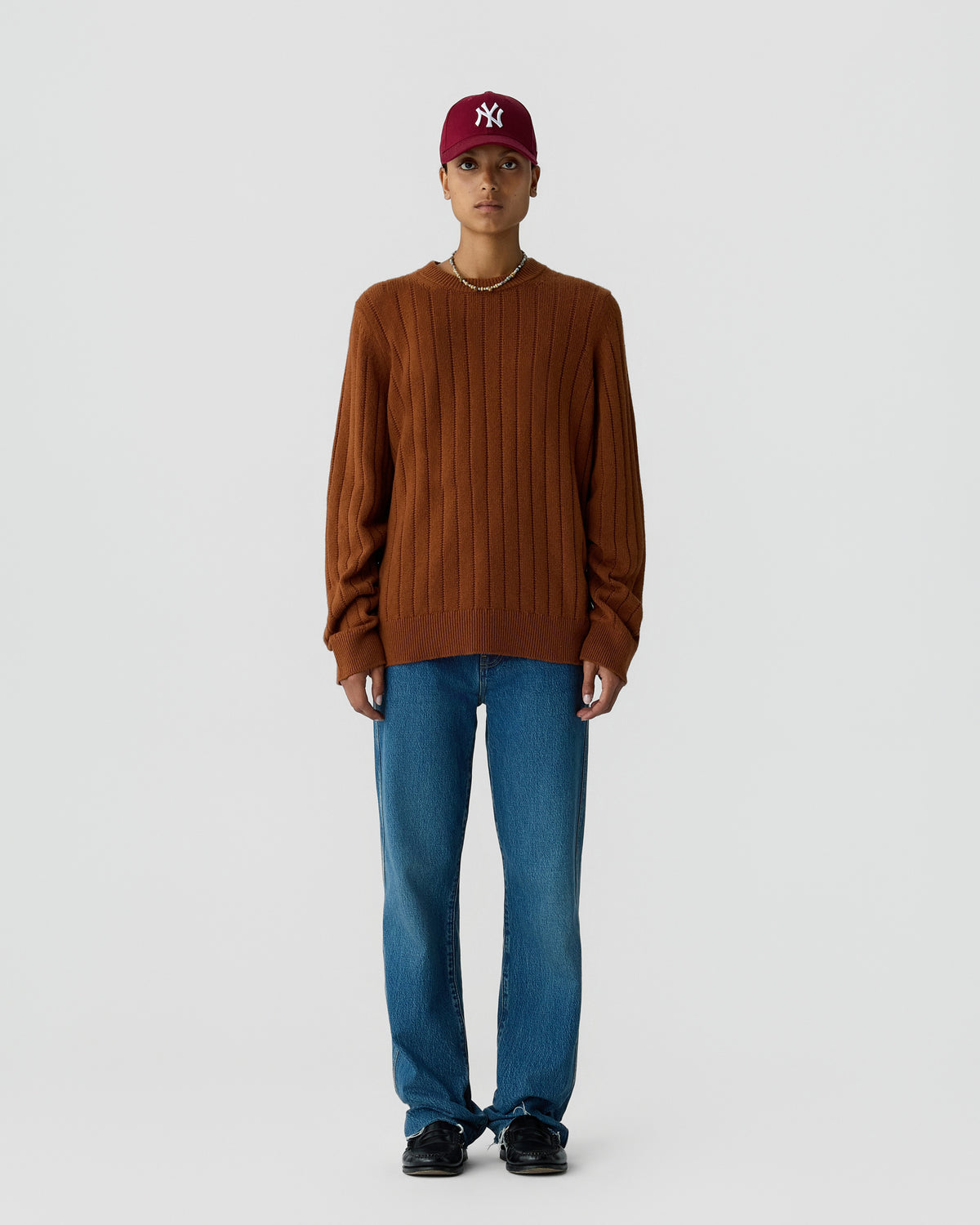 Young person wearing a brown ribbed sweater, blue jeans, black shoes, a red baseball cap, and a necklace, standing against a plain light gray background.