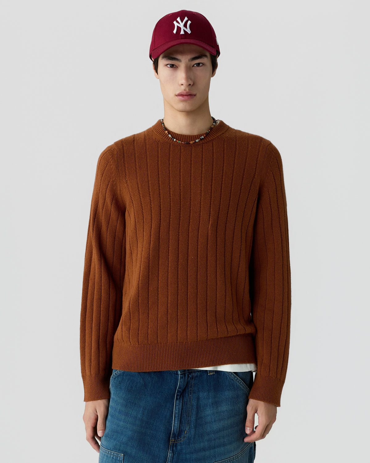 Young man wearing a ribbed brown sweater and blue jeans with a maroon baseball cap against a plain light gray background