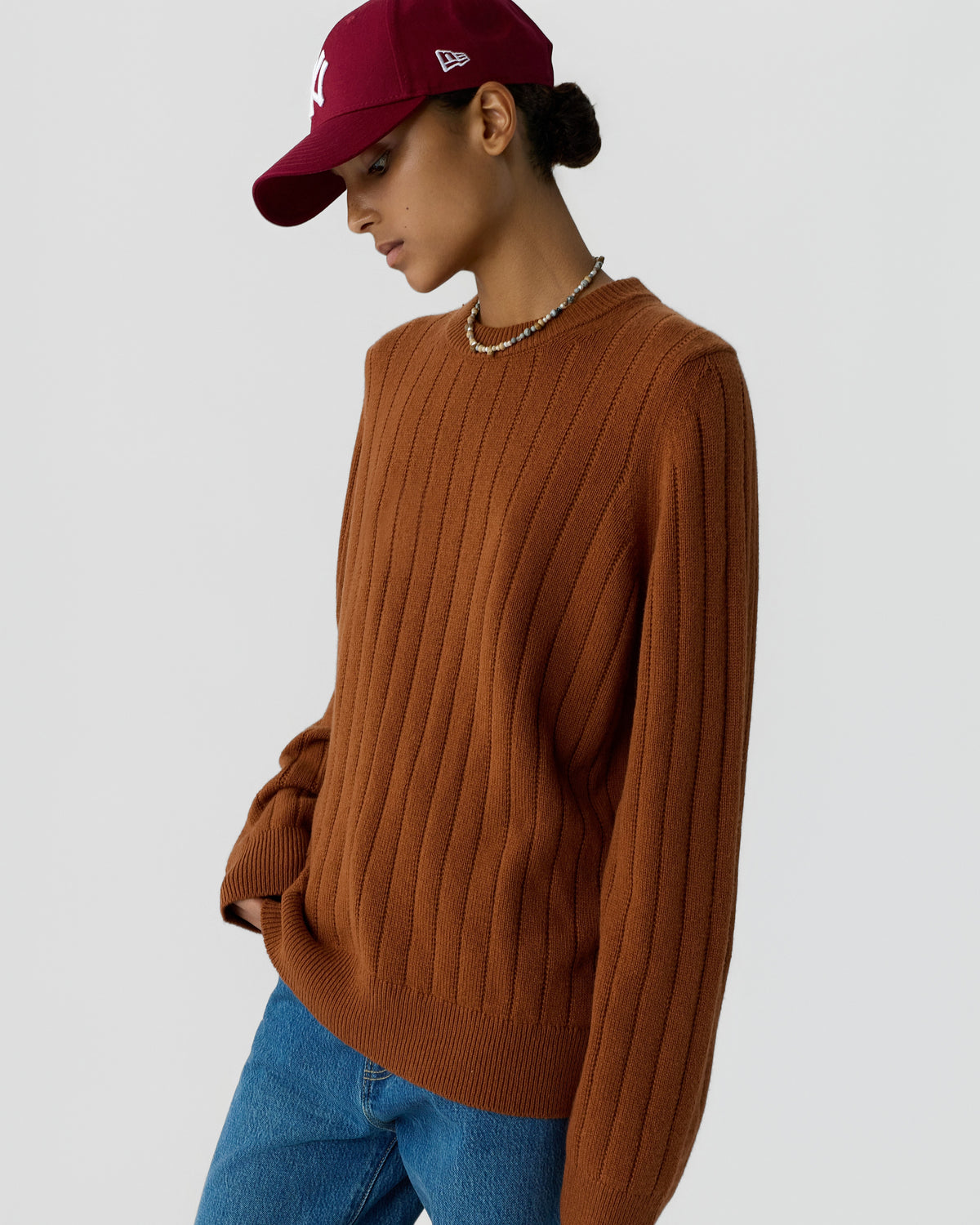 Woman wearing a rust-colored ribbed cashmere sweater, blue jeans, a maroon baseball cap, and a beaded necklace against a plain gray background.