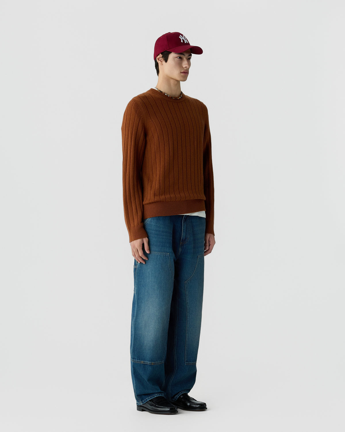 A man stands against a plain light gray background wearing a brown ribbed sweater, wide-legged blue jeans, black shoes, and a red baseball cap.