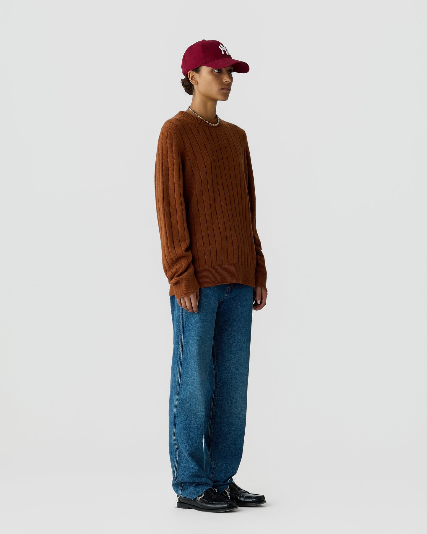 Person wearing a brown ribbed sweater, blue wide-leg jeans, black shoes, and a maroon baseball cap standing against a plain light gray background.