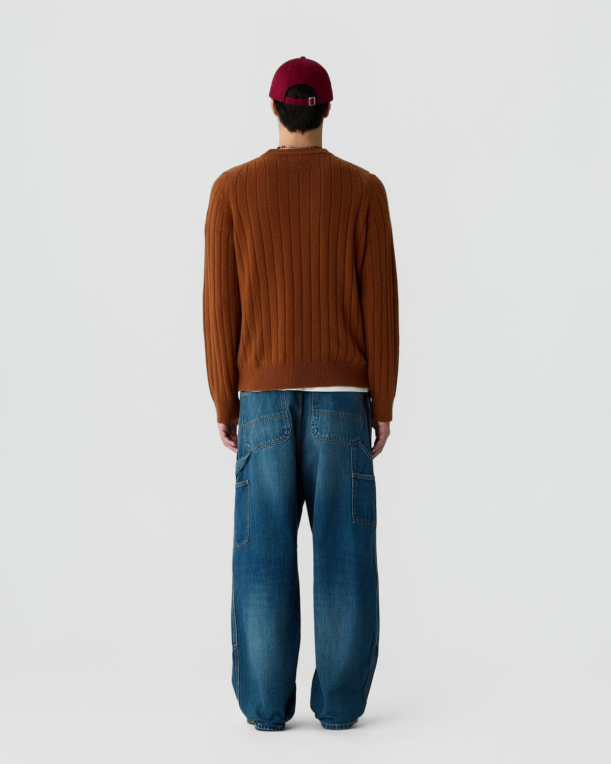Man wearing a rust-colored ribbed sweater, blue wide-leg jeans with multiple pockets, and a red baseball cap, standing facing away against a plain background.