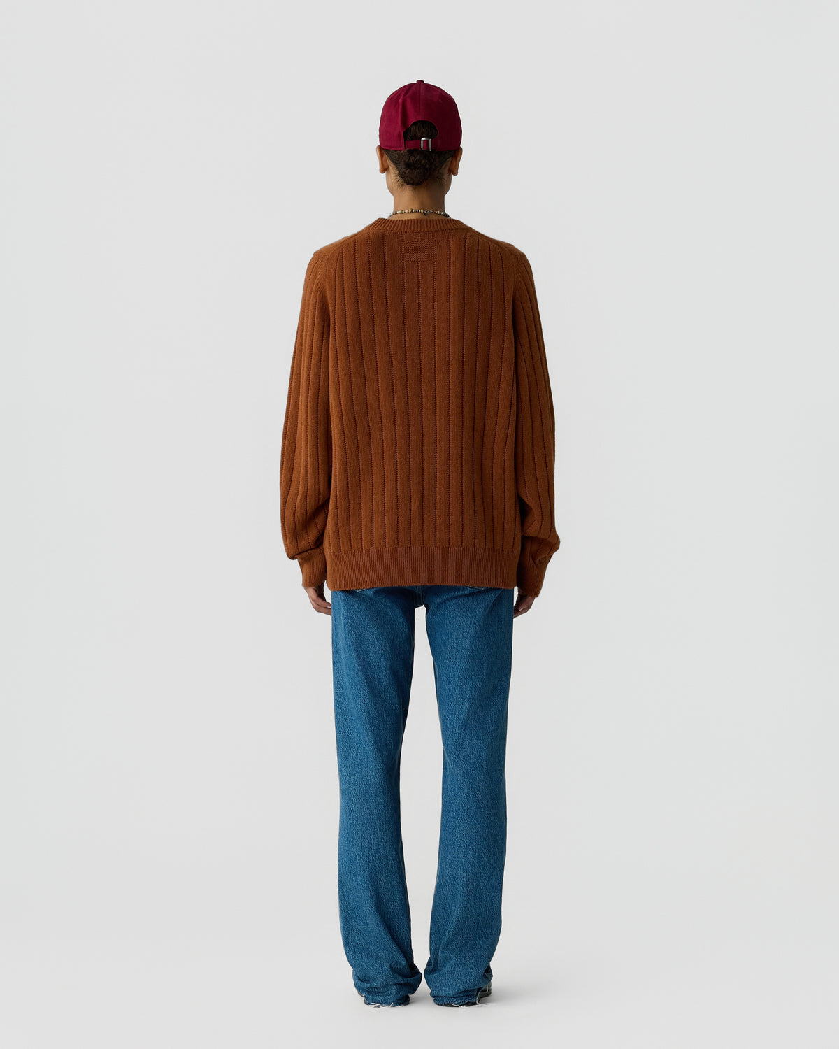 Person wearing a ribbed brown sweater and blue jeans with a red cap facing away against a plain light gray background