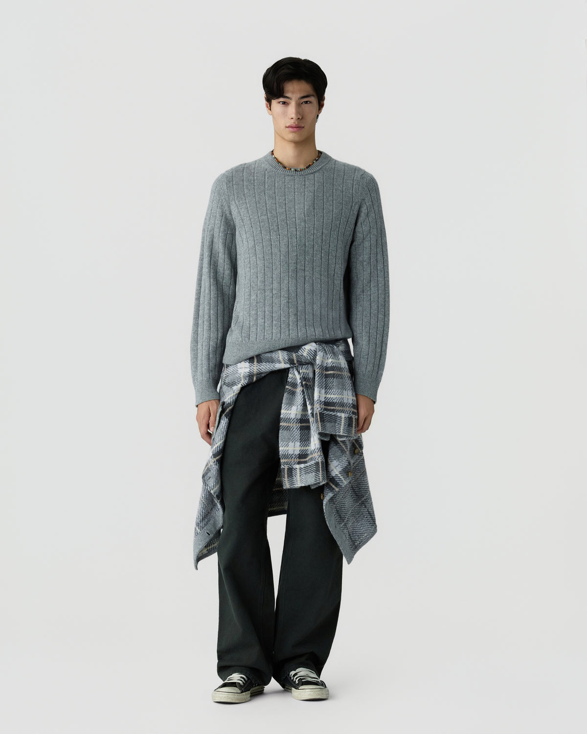 Man wearing a gray ribbed sweater with a plaid shirt tied around the waist, black wide-leg pants, and black and white sneakers, standing against a plain white background.