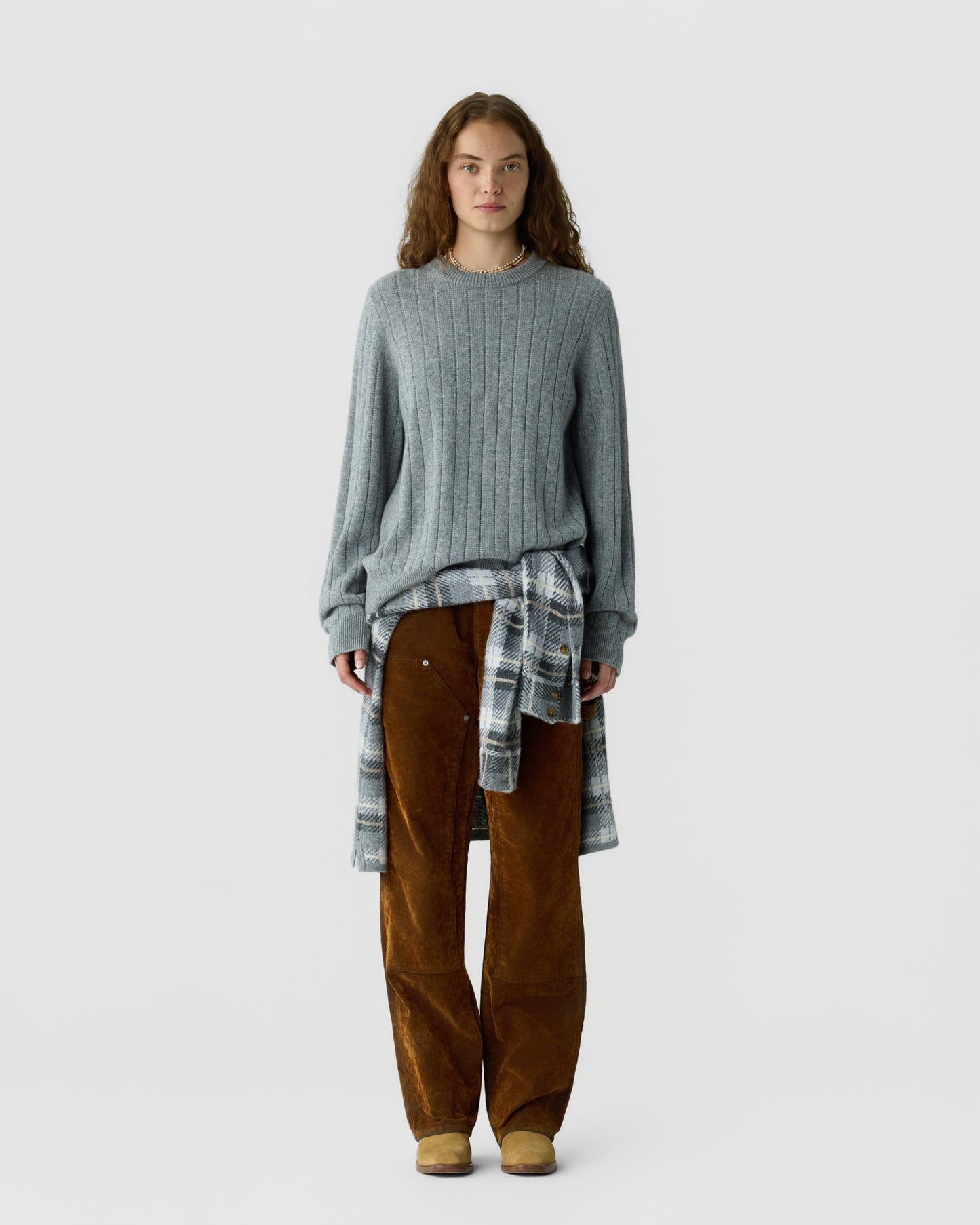 Young woman standing against a plain background wearing a gray ribbed sweater, brown corduroy pants, and a plaid shirt tied around her waist.