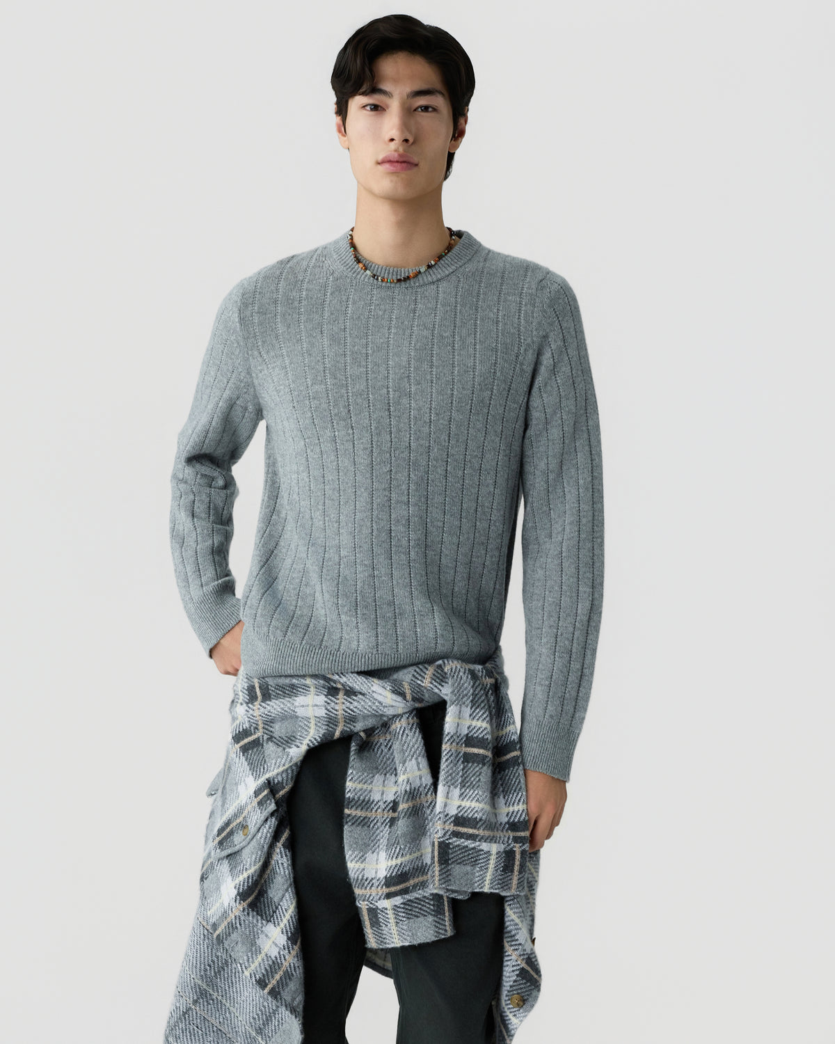 Young man wearing a light gray ribbed knit sweater with a plaid shirt tied around his waist and a beaded necklace against a plain white background.