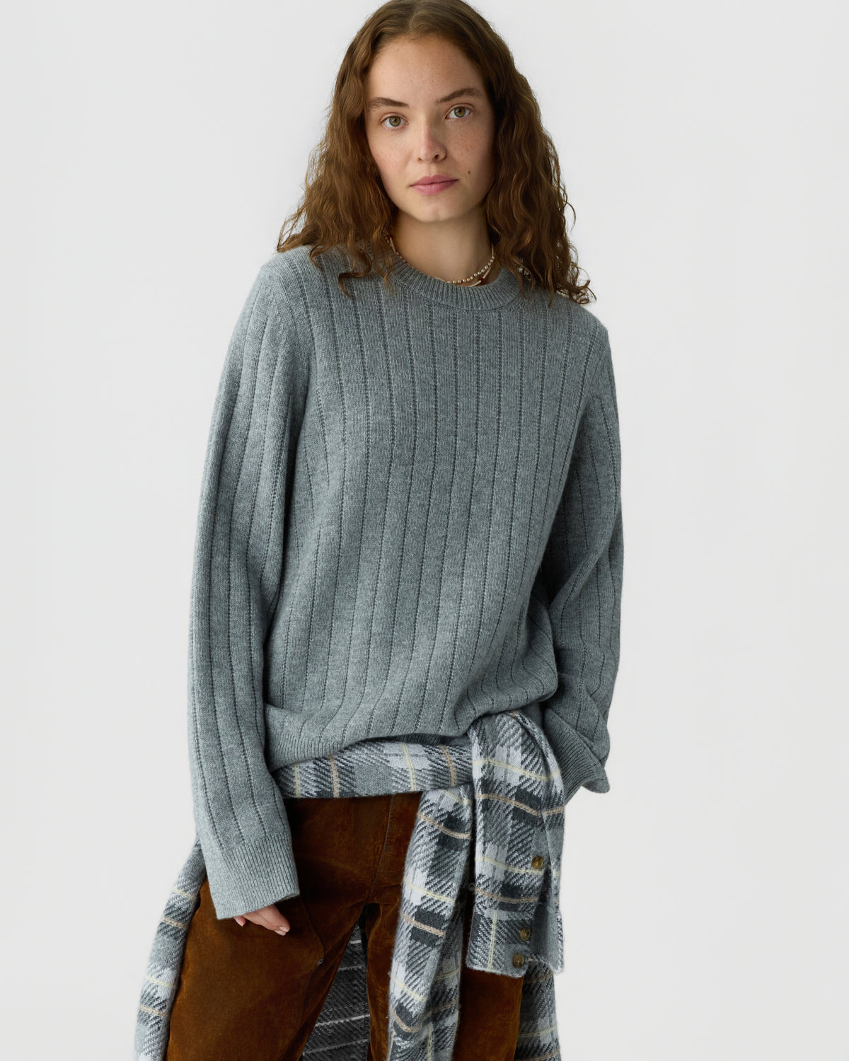 Young woman with long curly hair wearing a gray ribbed sweater and brown pants, with a plaid shirt tied around her waist, standing against a plain light background.
