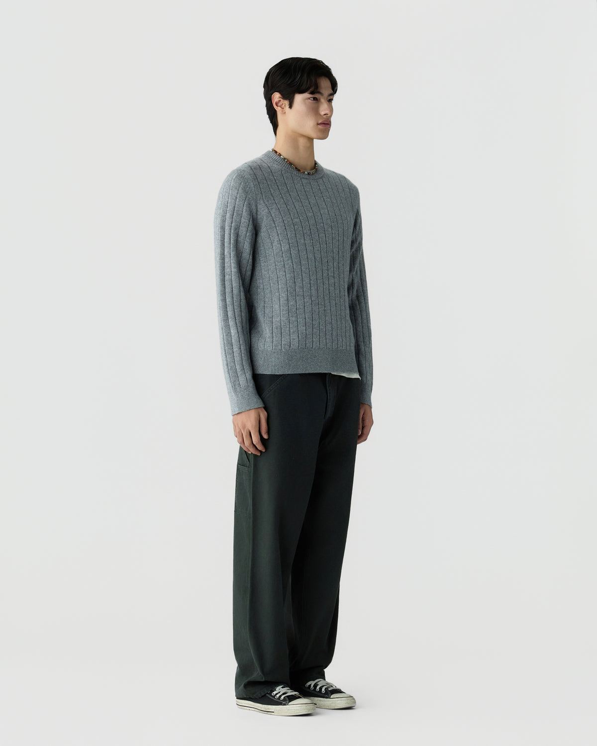 Young man standing against plain background wearing a gray ribbed sweater, dark wide-leg pants, and black sneakers with white soles.