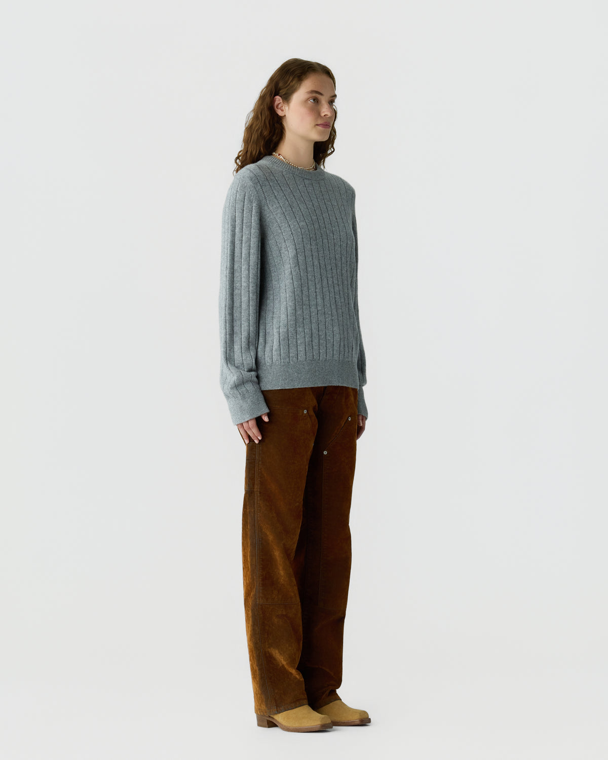 Woman with curly brown hair wears a gray ribbed sweater and loose brown corduroy pants, standing sideways against a plain light background.