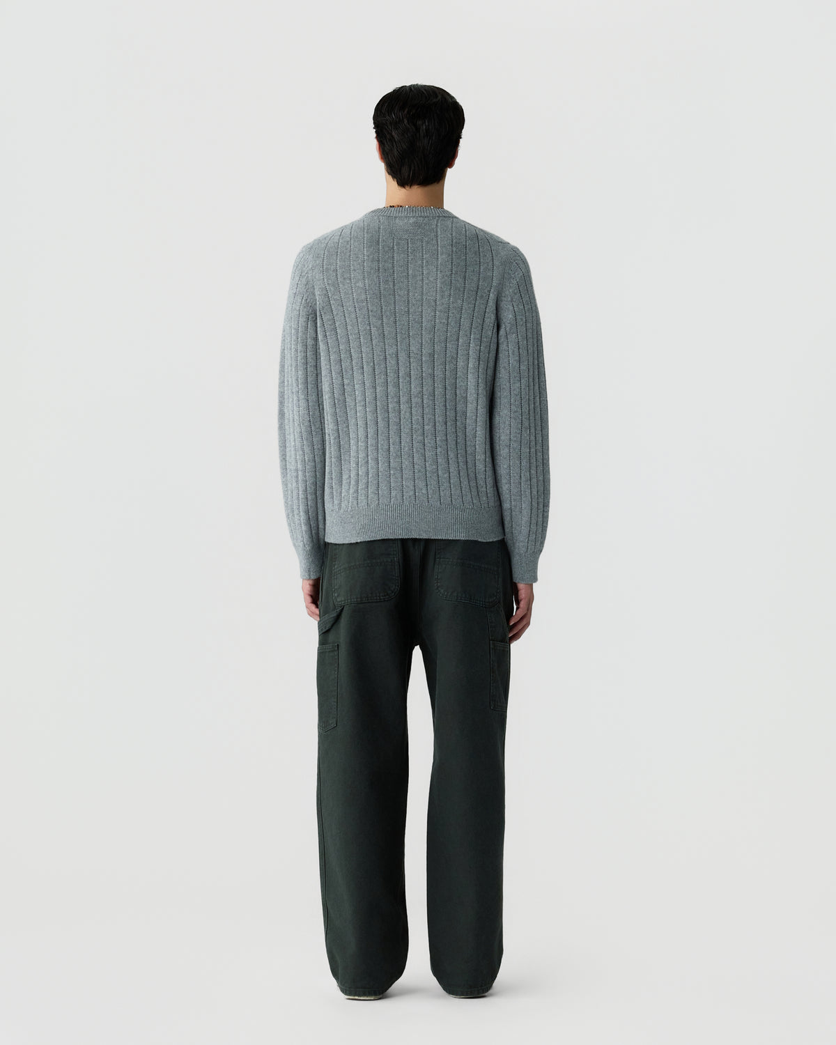 A person standing with their back to the camera wearing a light gray ribbed sweater and loose dark pants against a plain white background.