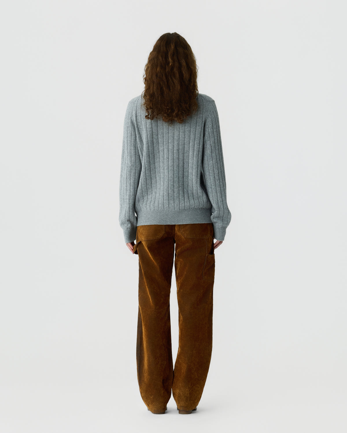 Person with long curly hair wearing a light gray ribbed sweater and loose-fitting brown corduroy pants standing facing away against a plain white background.