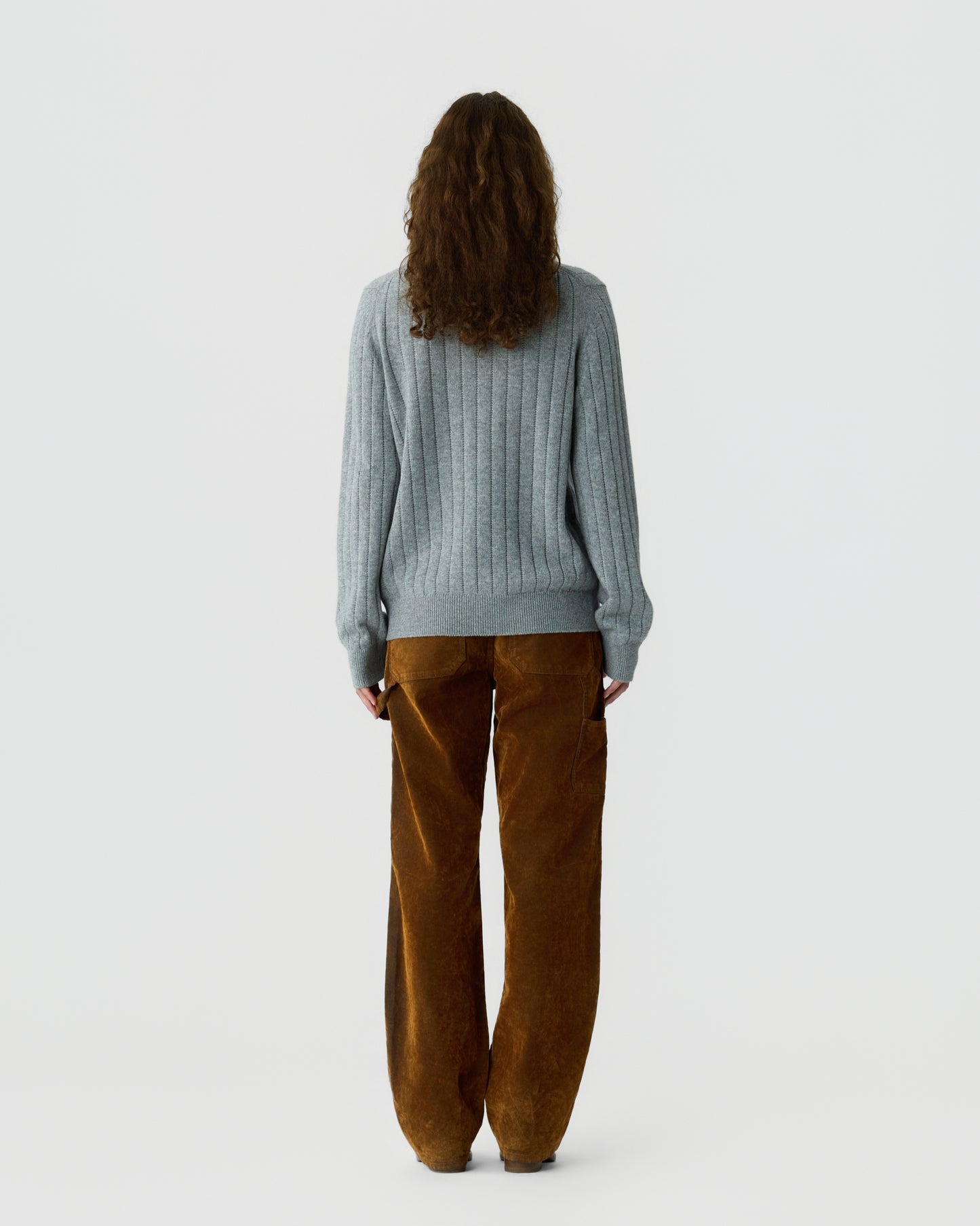 Person with long curly hair wearing a light gray ribbed sweater and loose-fitting brown corduroy pants standing facing away against a plain white background.