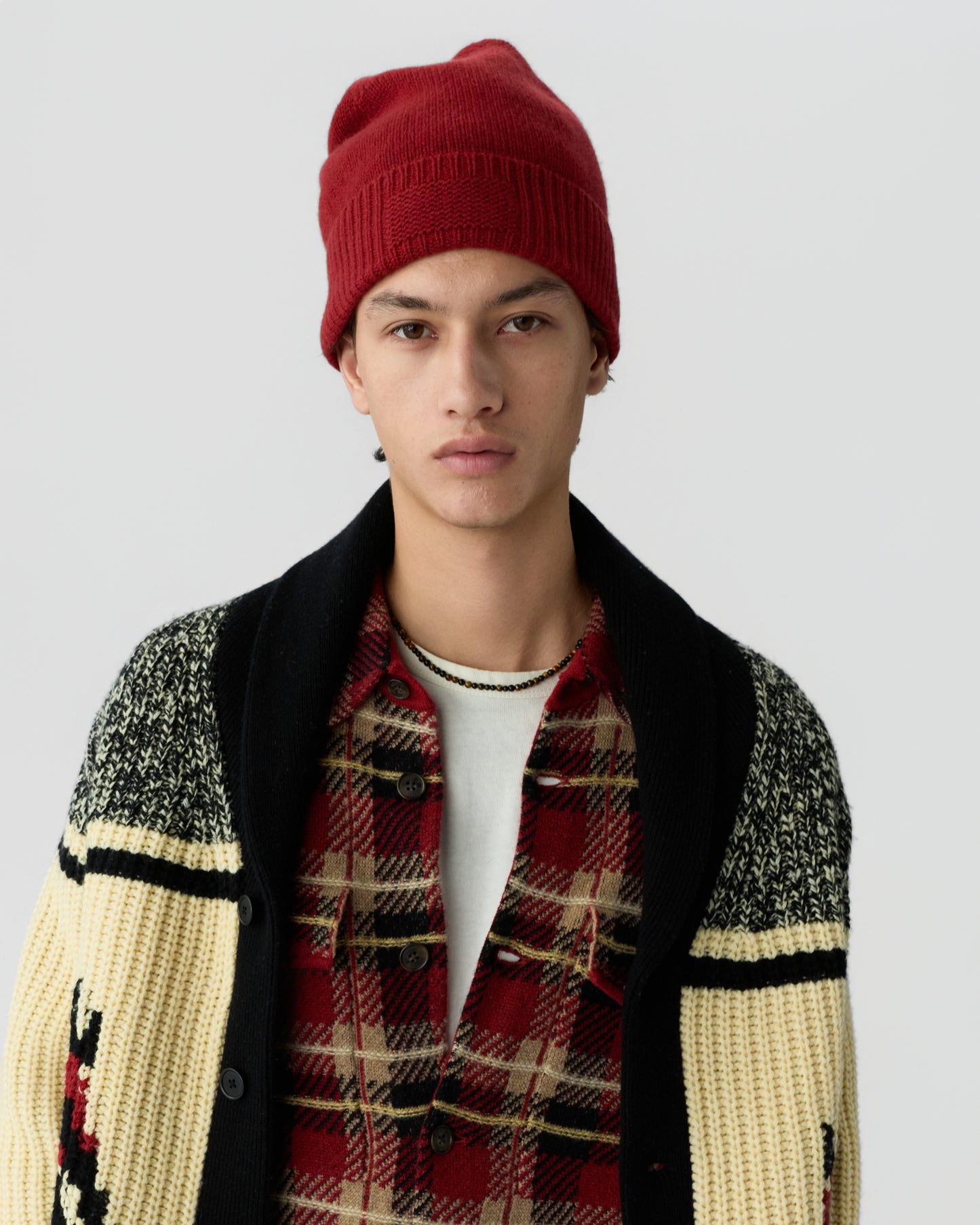 Young man wearing a red beanie, a red and black plaid shirt layered over a white undershirt, and a chunky knit cardigan with cream, black, and red accents against a plain background.