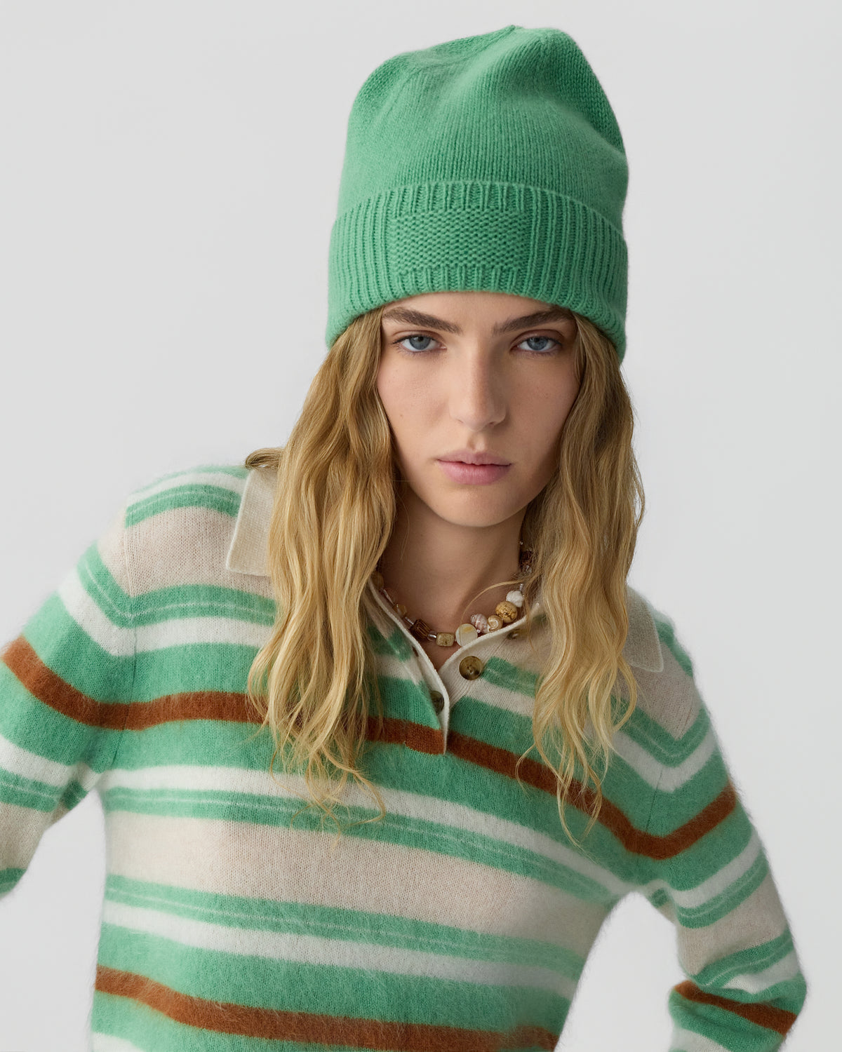Young woman with long blonde hair wearing a green cashmere beanie and a striped green, beige, and brown cashmere sweater against a plain gray background.