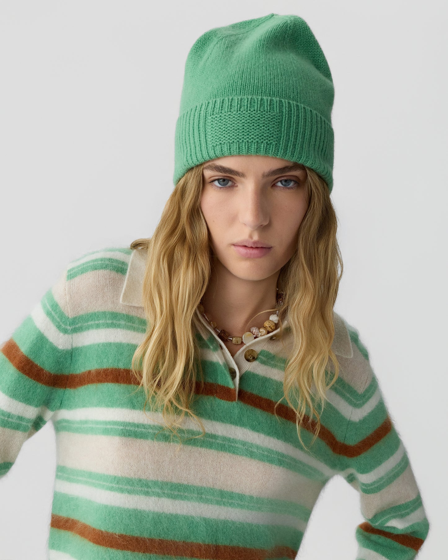 Young woman with long blonde hair wearing a green cashmere beanie and a striped green, beige, and brown cashmere sweater against a plain gray background.