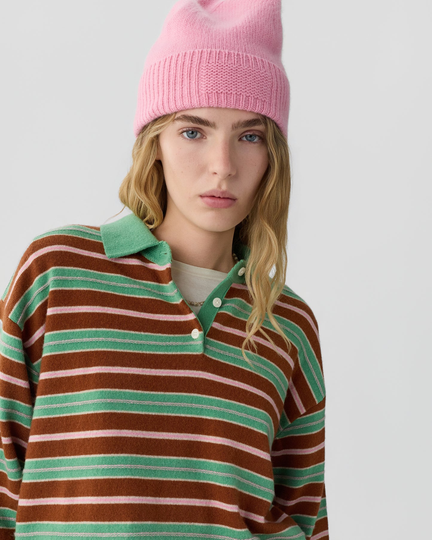 Young woman with long blonde hair wearing a pink knit beanie and a brown and green striped long-sleeve shirt over a light-colored top, looking at the camera.