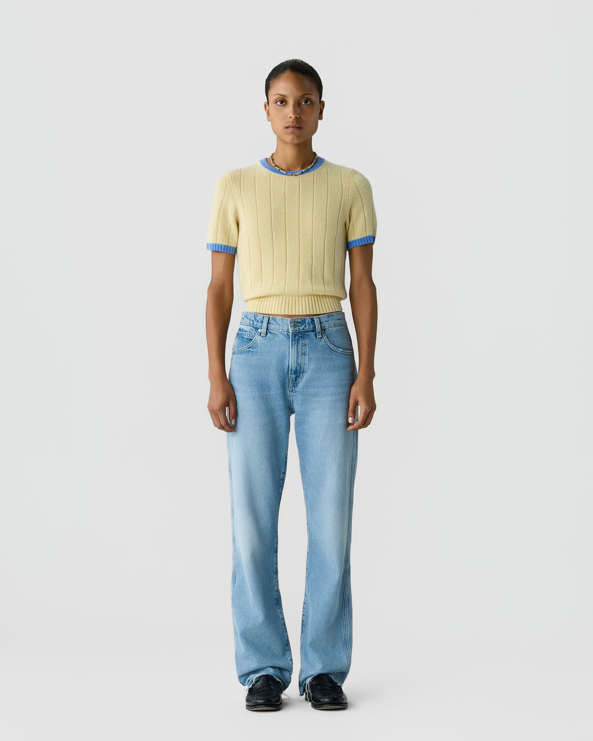 Model wearing a pale yellow ribbed short sleeve knit top with blue trim blue jeans and black shoes against a light gray background