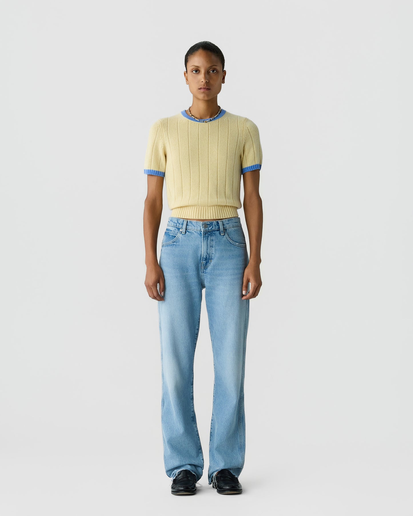 Model wearing a pale yellow ribbed short sleeve knit top with blue trim blue jeans and black shoes against a light gray background