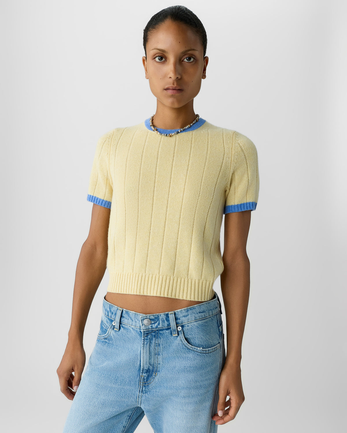 Model wearing a pale yellow ribbed short sleeve knit sweater with blue trim and light wash high waisted jeans against a plain gray background