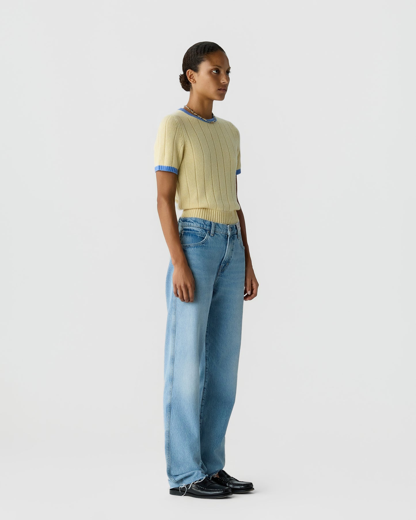 Full length view of a model wearing a pale yellow ribbed short sleeve top with blue trim blue jeans and black shoes against a light gray background