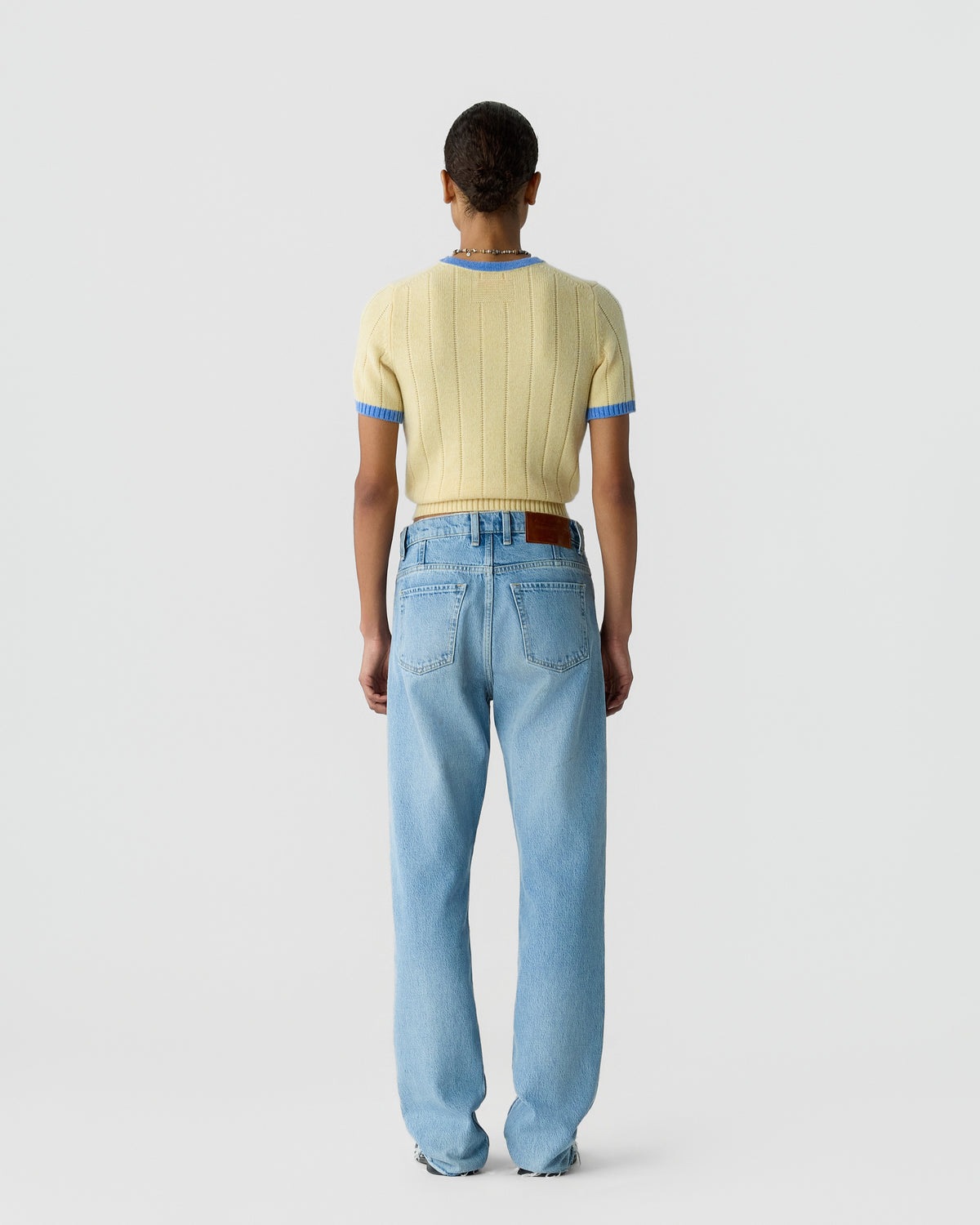 Person wearing a short-sleeve, light yellow knit sweater with blue trim and light blue straight-leg jeans, standing facing away against a plain background.