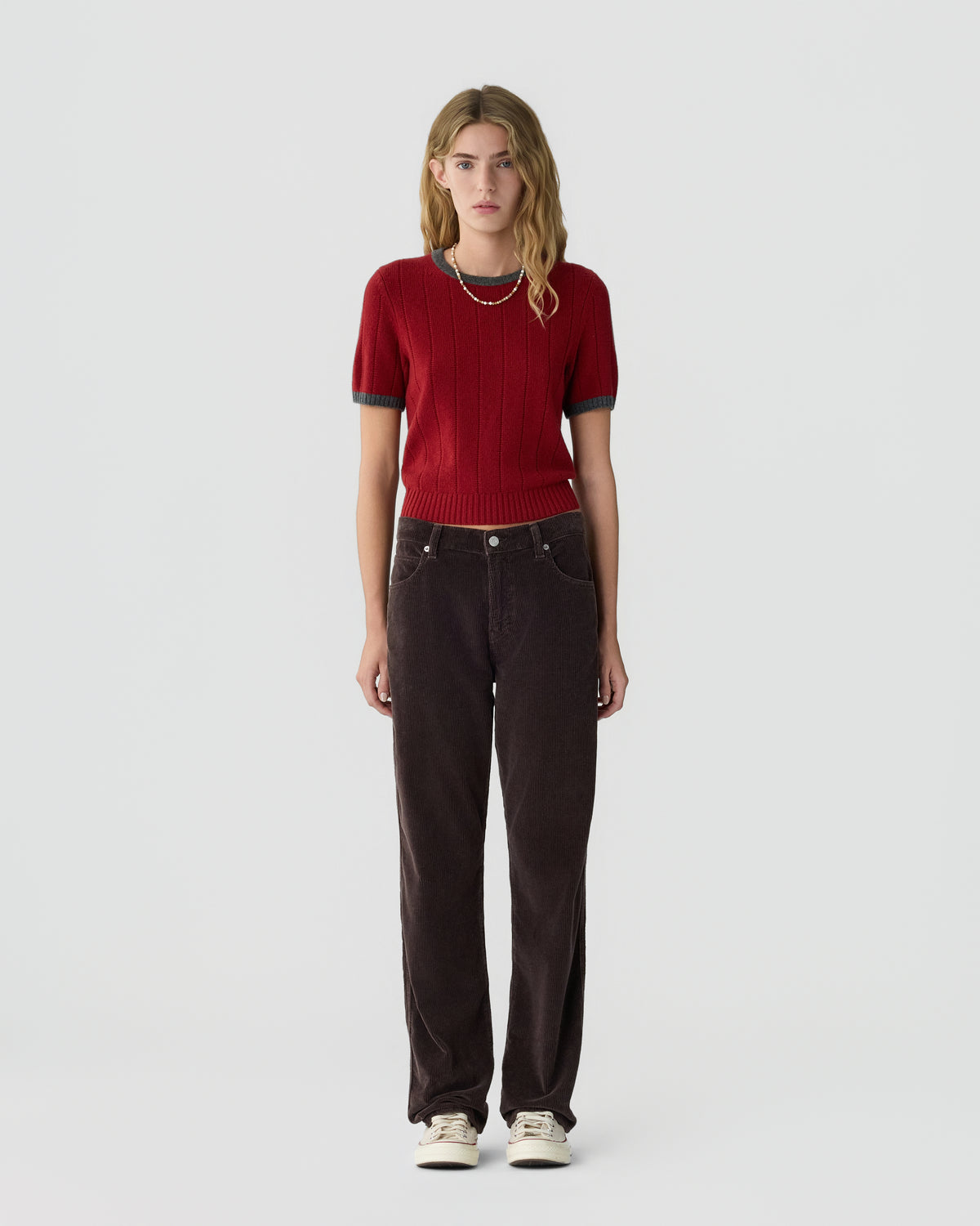 Young woman with blonde wavy hair wearing a red short-sleeve cashmere sweater paired with dark brown wide-leg pants and white sneakers, standing against a plain white background.